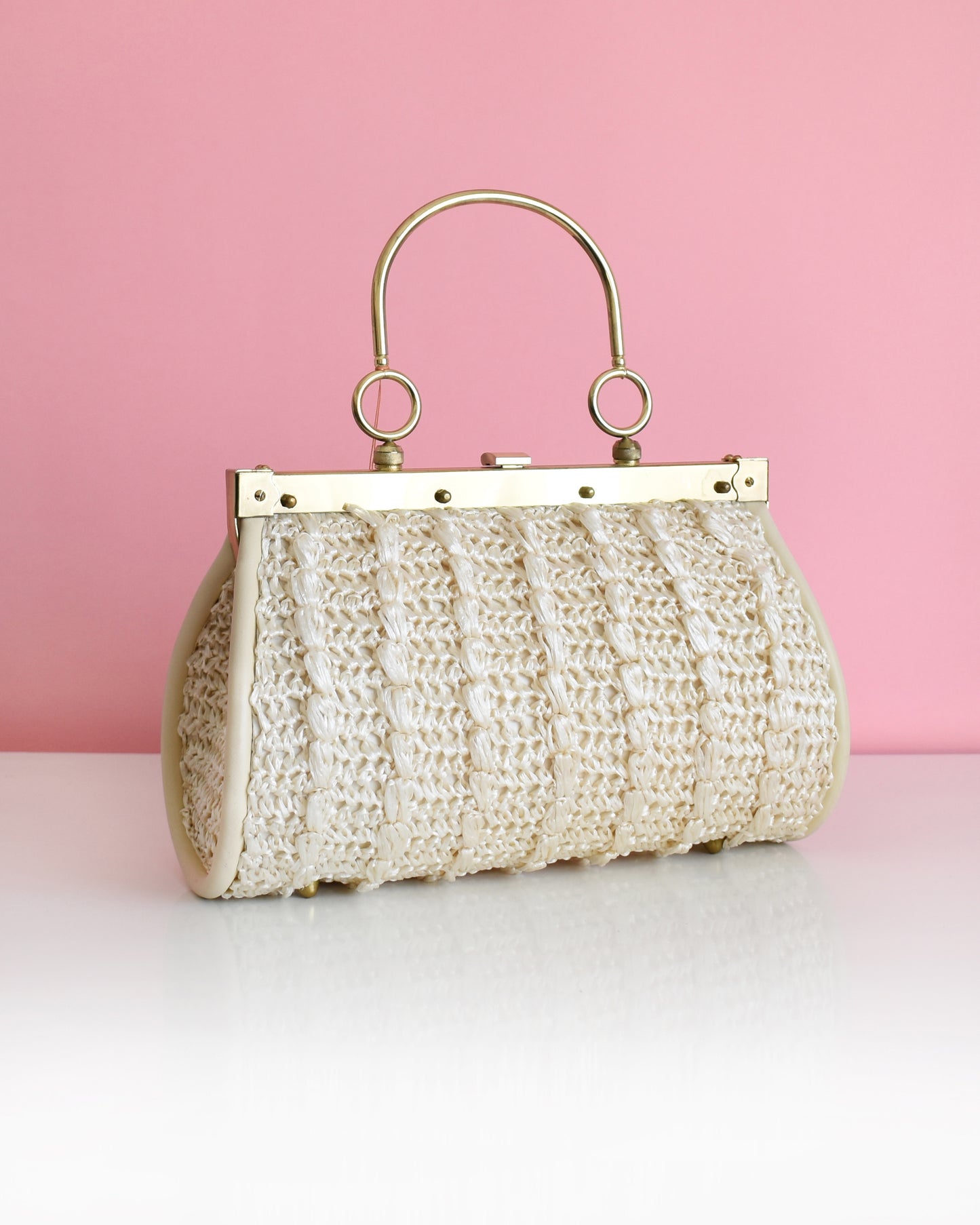 a vintage 1960s white woven handbag with gold frame and handle against a pink background