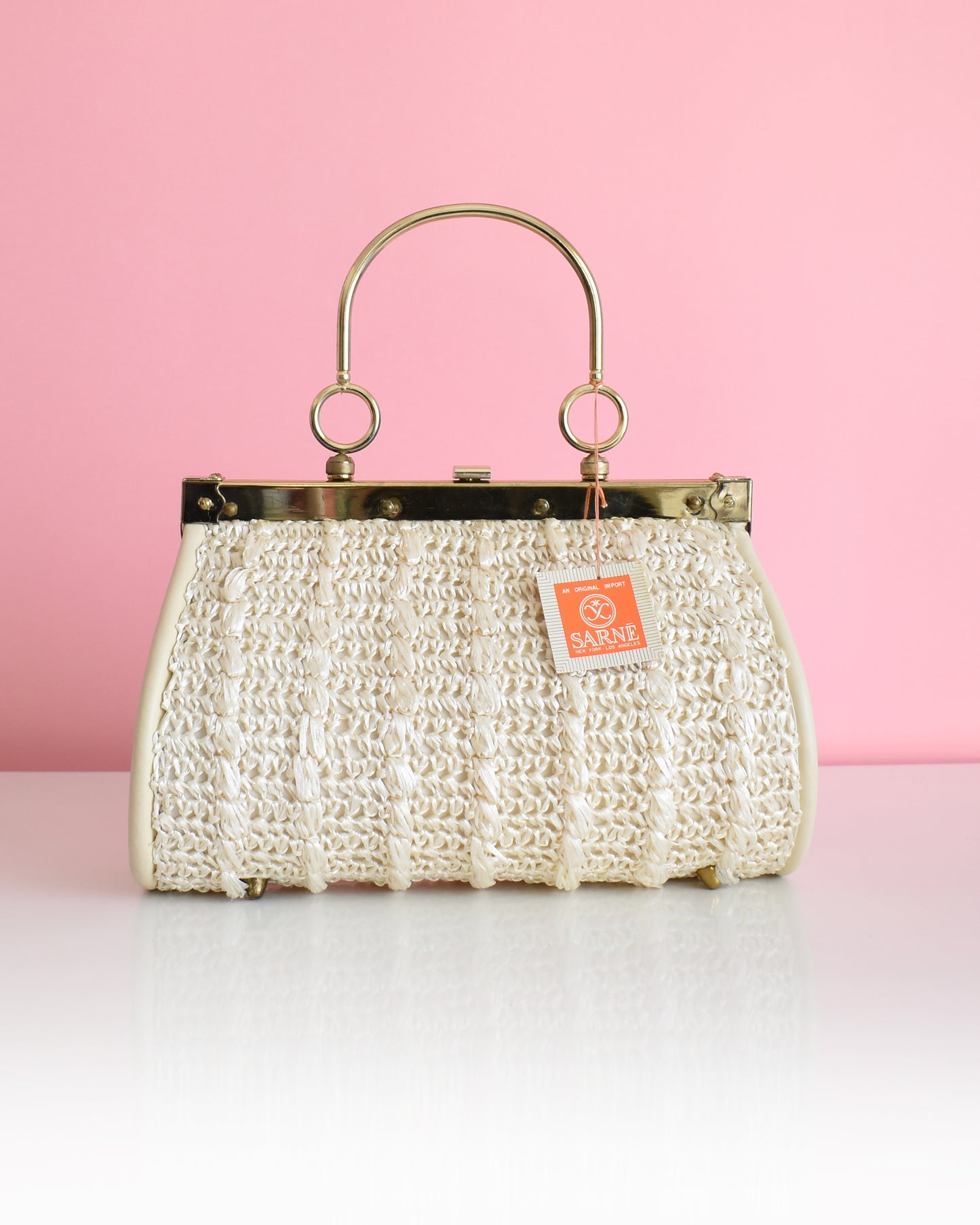 a vintage 1960s white woven handbag with gold frame and handle against a pink background