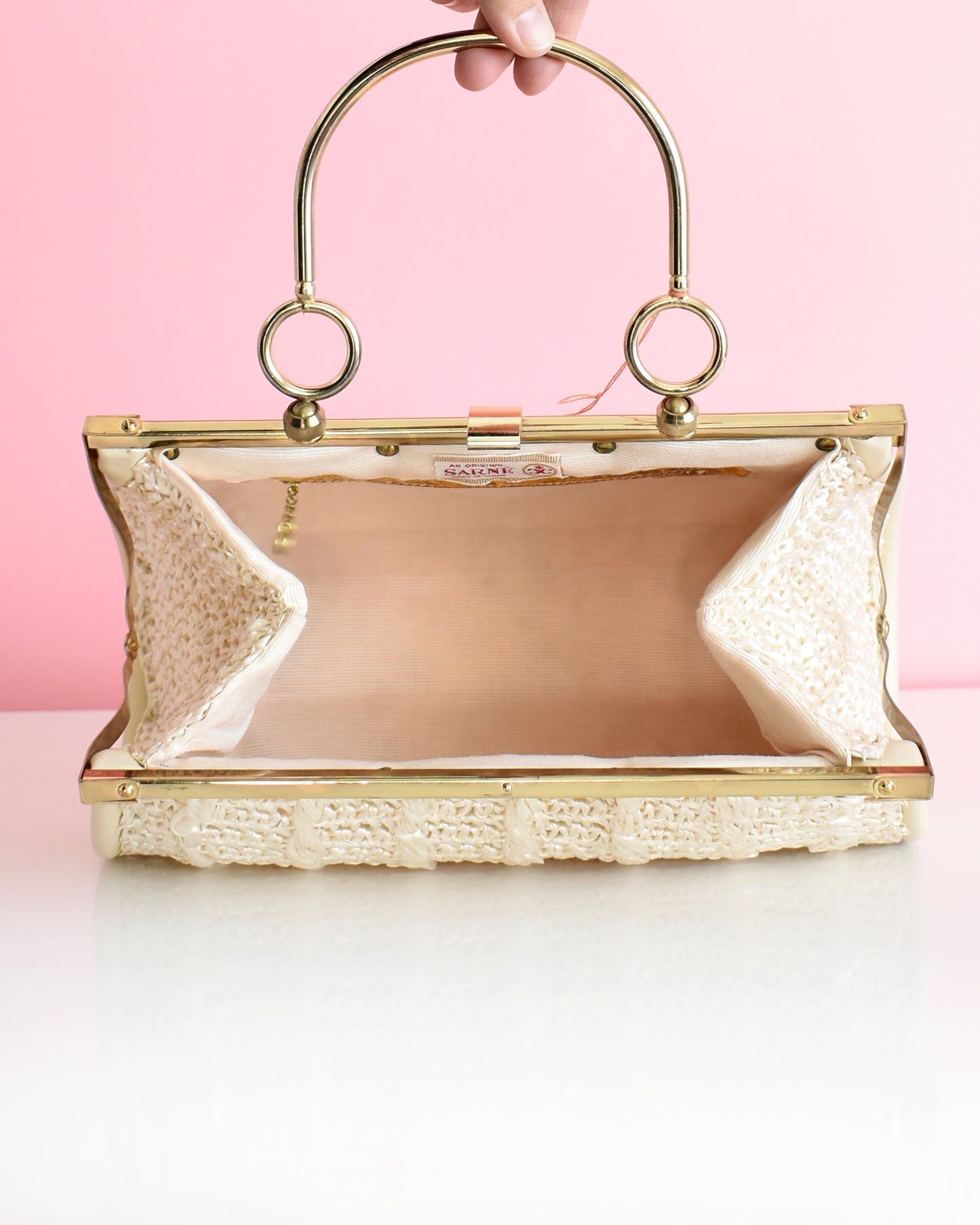 a vintage 1960s white woven handbag with gold frame and handle against a pink background which is open showing the inside lining.