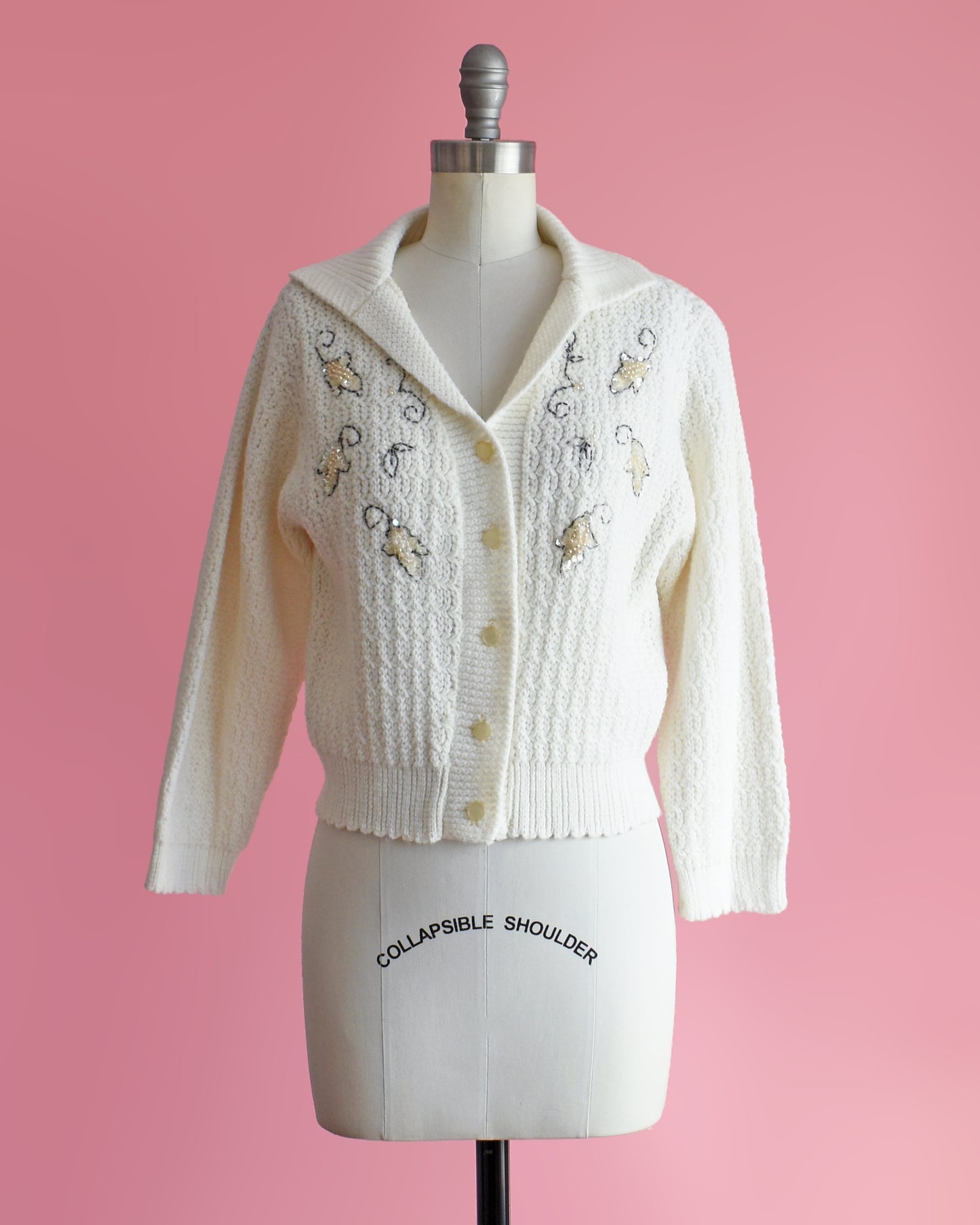 a vintage white cardigan with embroidered details on a mannequin against a pink background