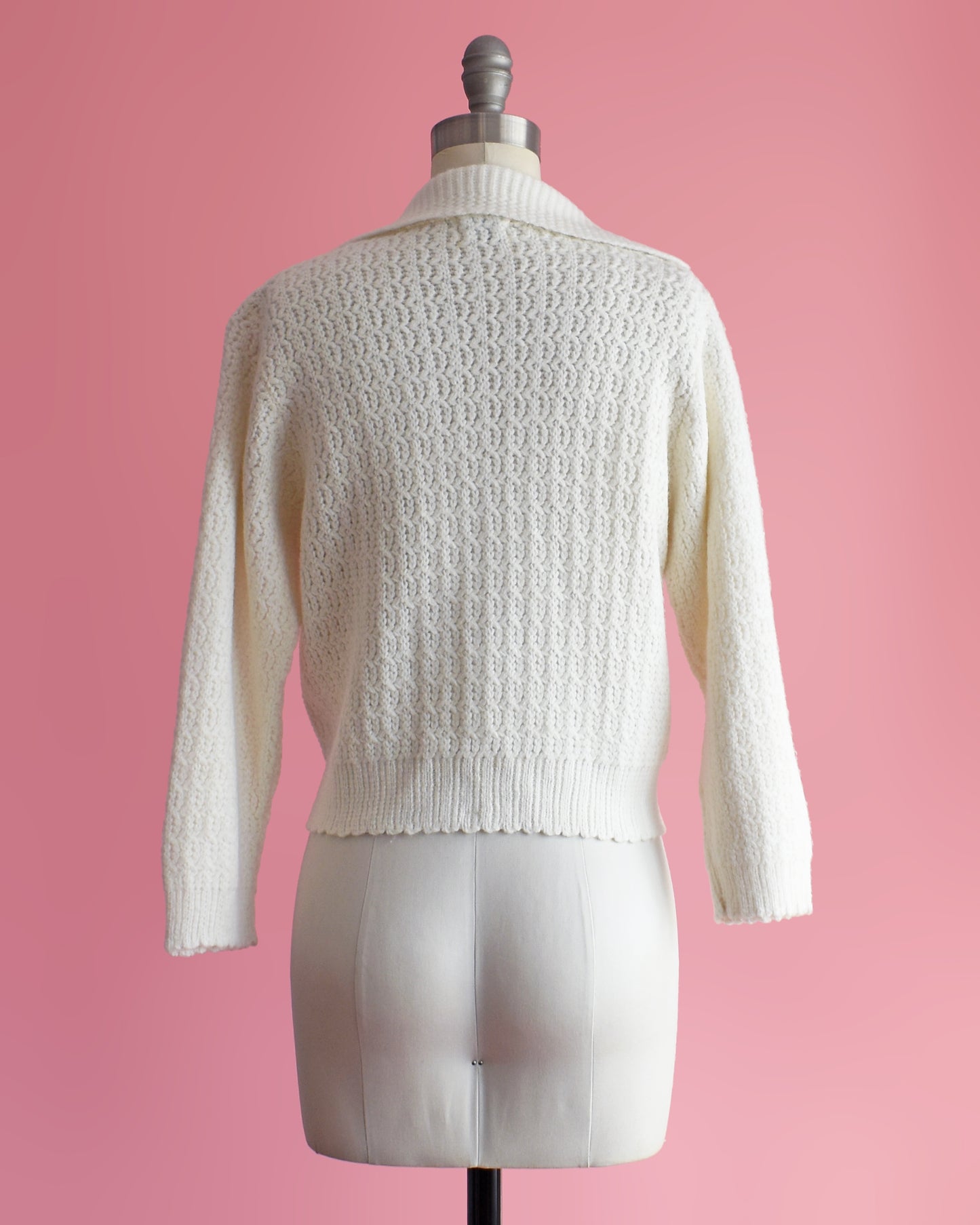 back view of a vintage white cardigan with embroidered details on a mannequin against a pink background