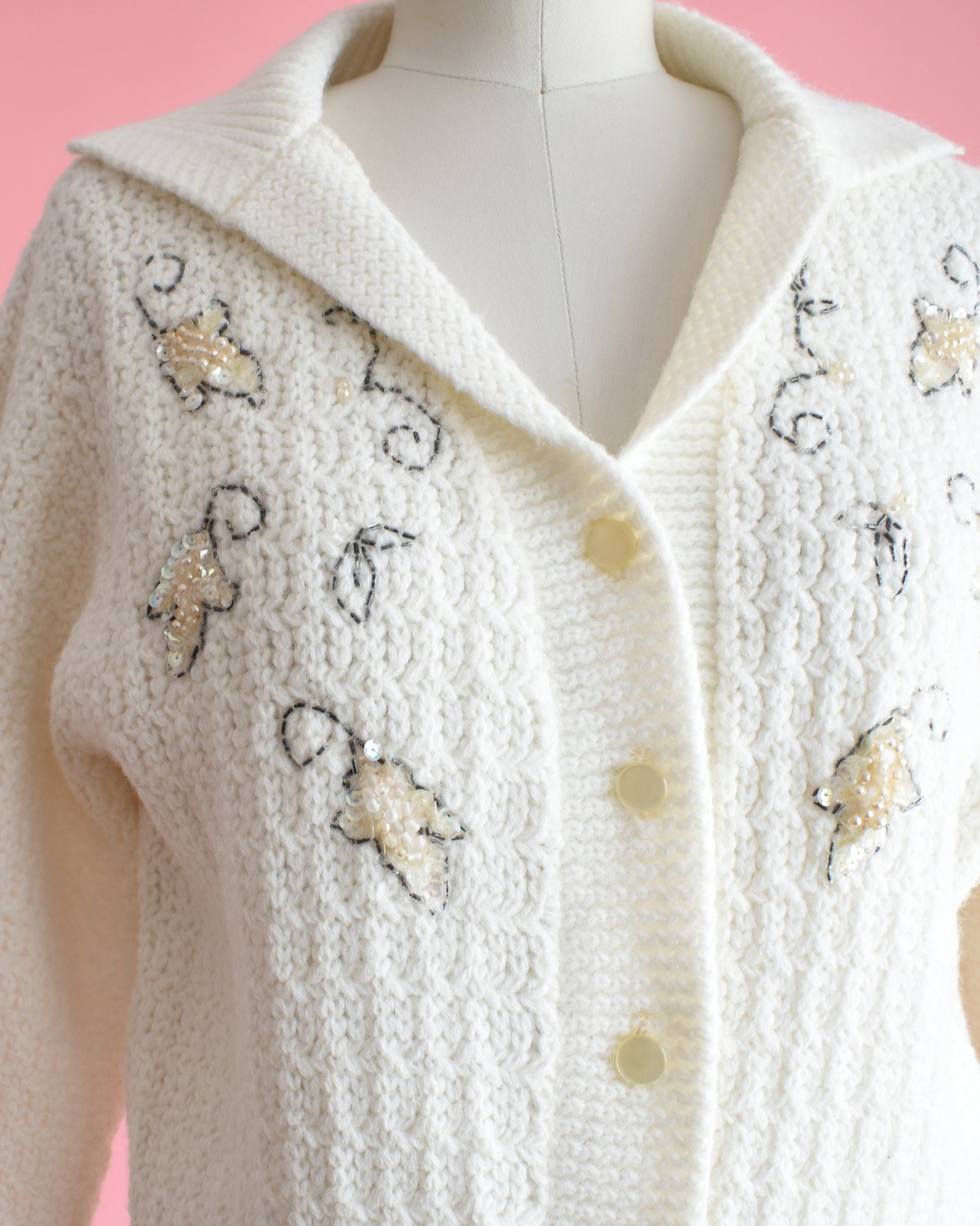 close up of a vintage white cardigan with embroidered details on a mannequin against a pink background