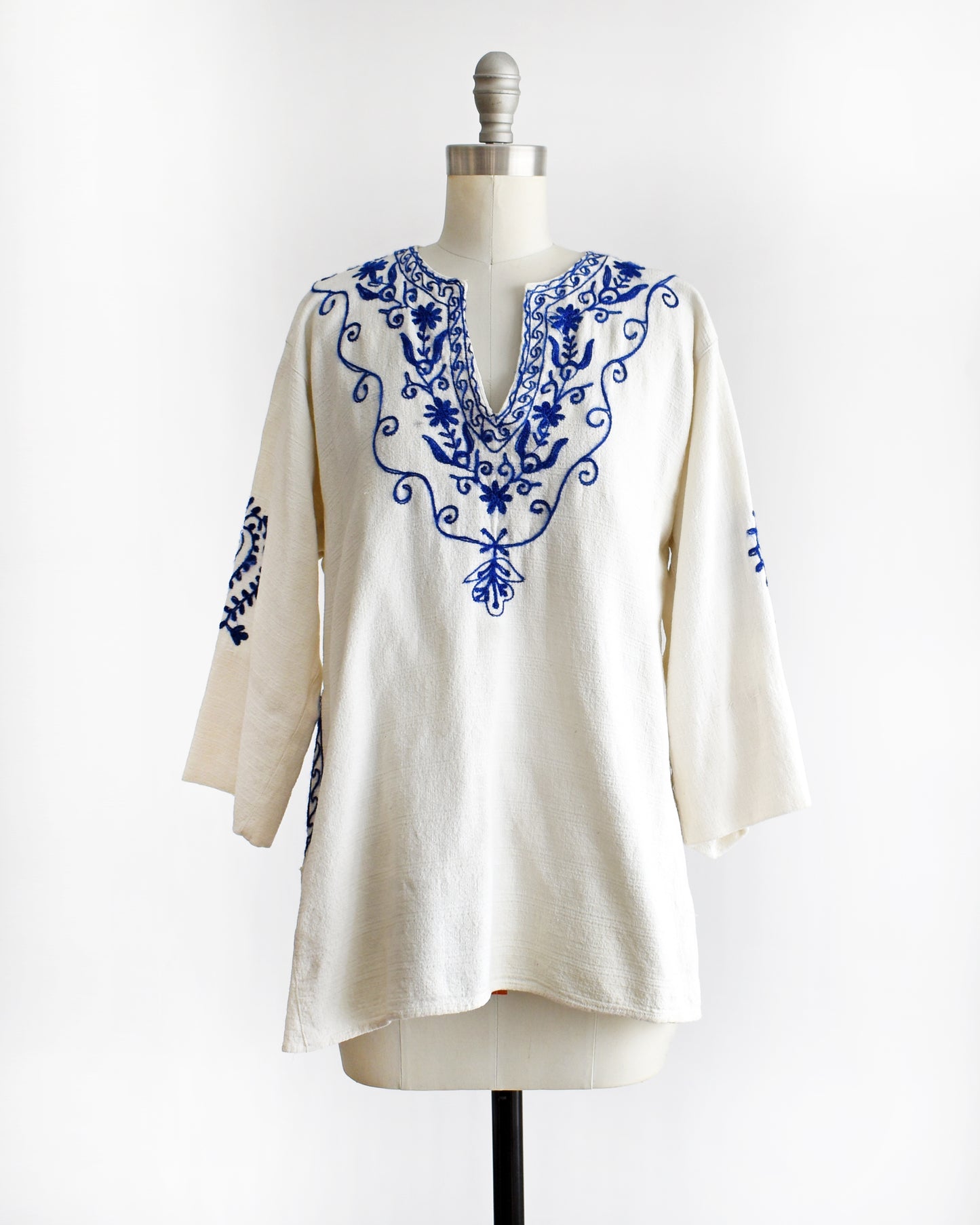 a vintage 1970s white blouse with blue embroidery on a mannequin against a white background