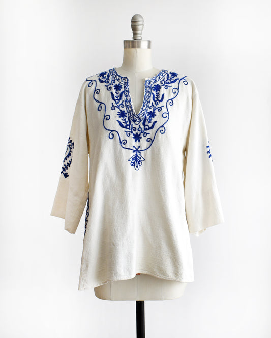 a vintage 1970s white blouse with blue embroidery on a mannequin against a white background