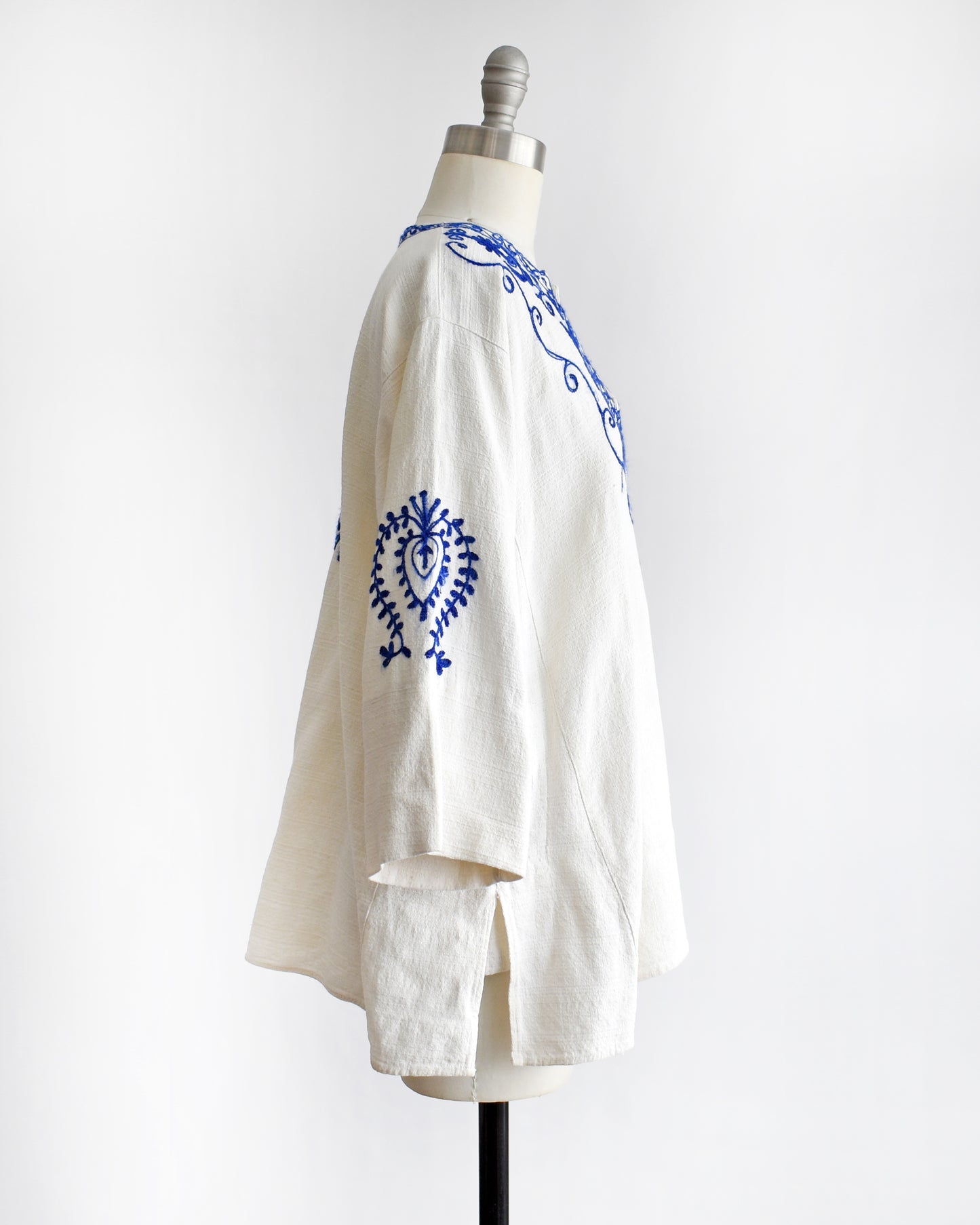 side view of a vintage 1970s white blouse with blue embroidery on a mannequin against a white background