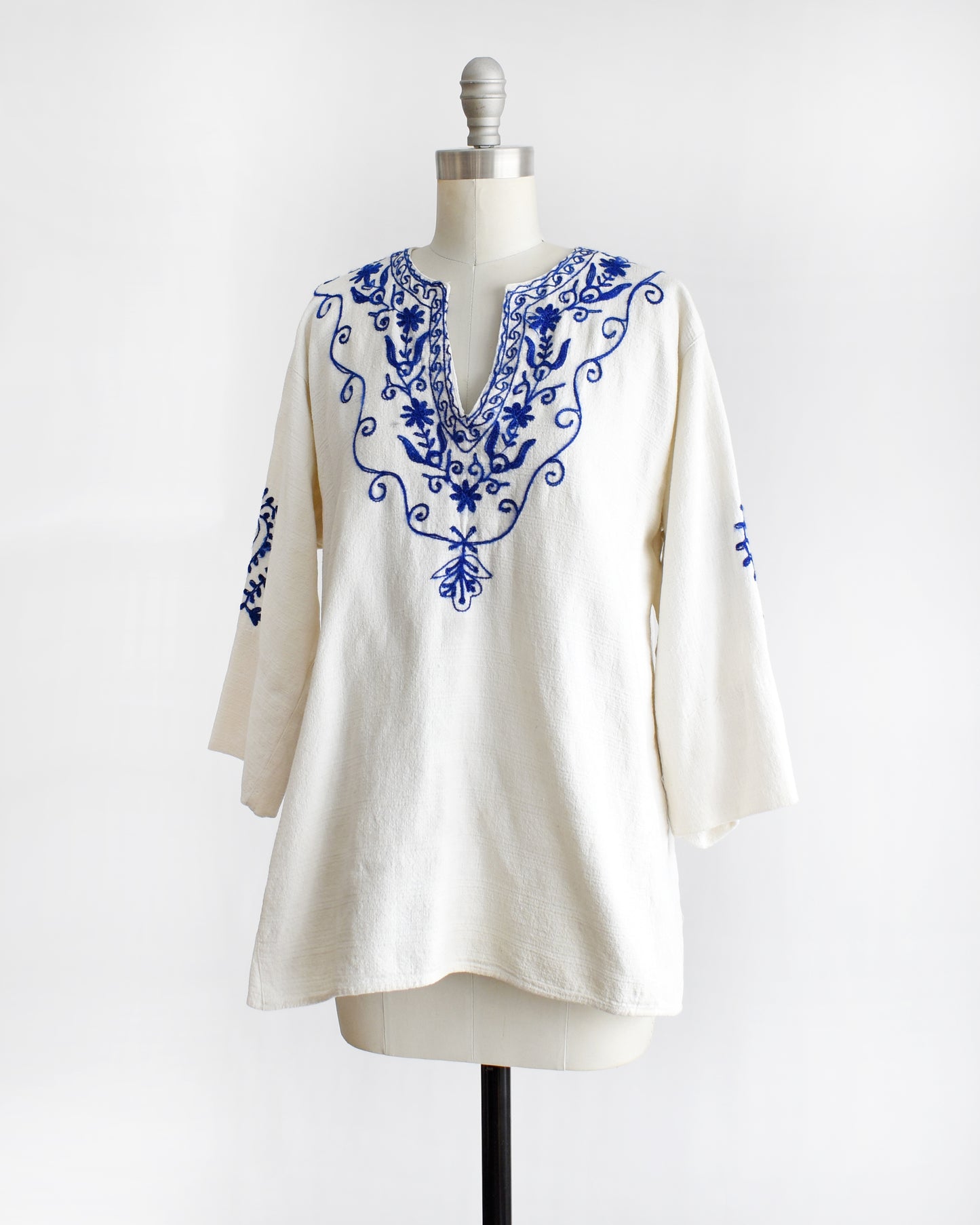 side front view of a vintage 1970s white blouse with blue embroidery on a mannequin against a white background