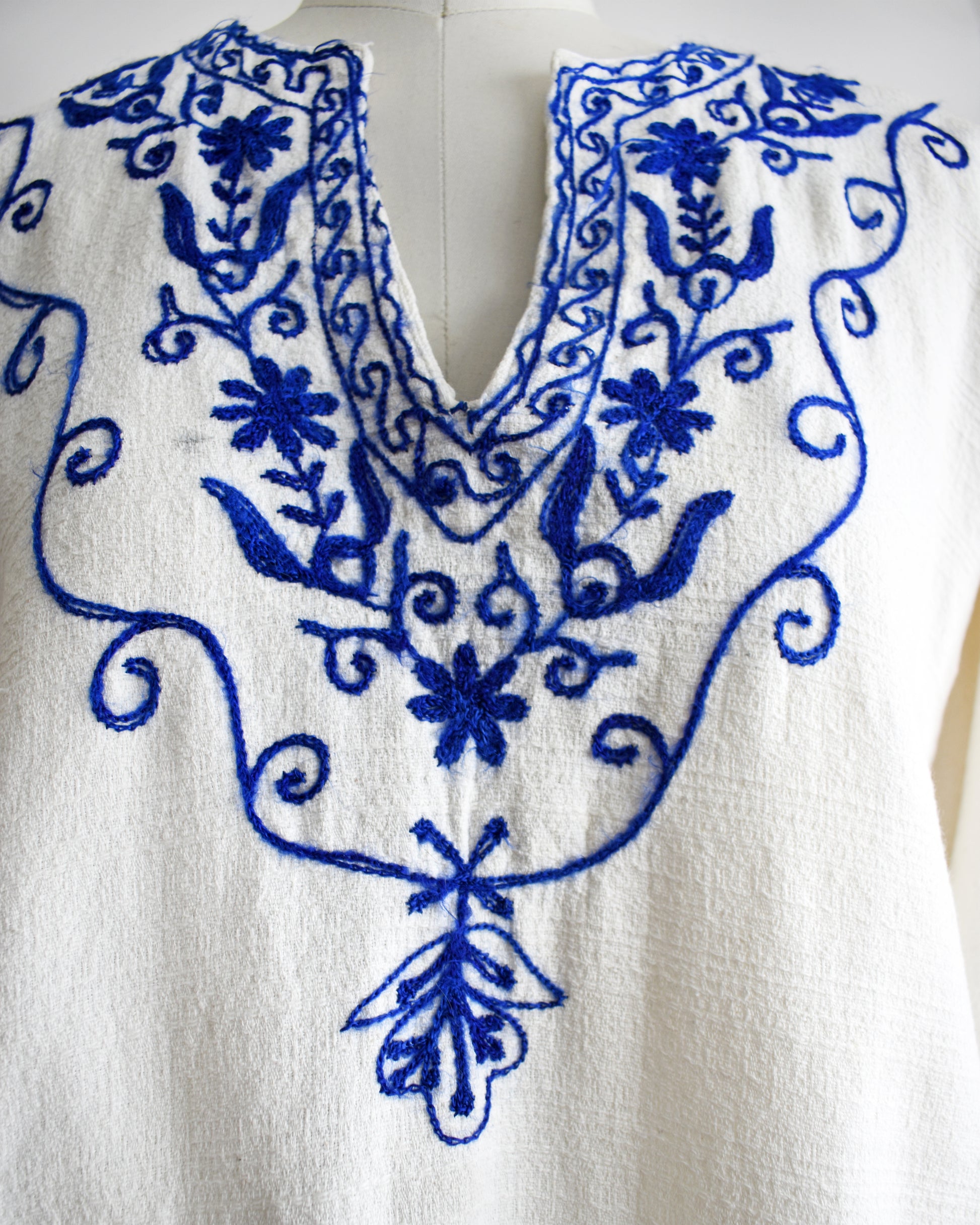 close up of the embroidered neckline on a vintage 1970s white blouse with blue embroidery on a mannequin against a white background