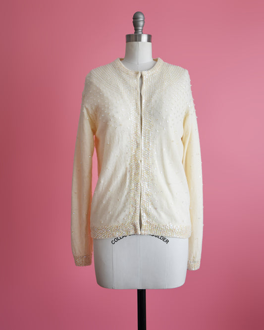 A vintage cream white wool cardigan that has iridescent sequin detail. The cardigan is on a dress form set on a pink background.