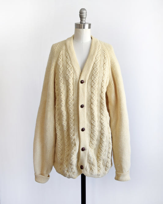 A vintage cream cable knit cardigan with dark buttons down the front. The cardigan is modeled on a dress form.