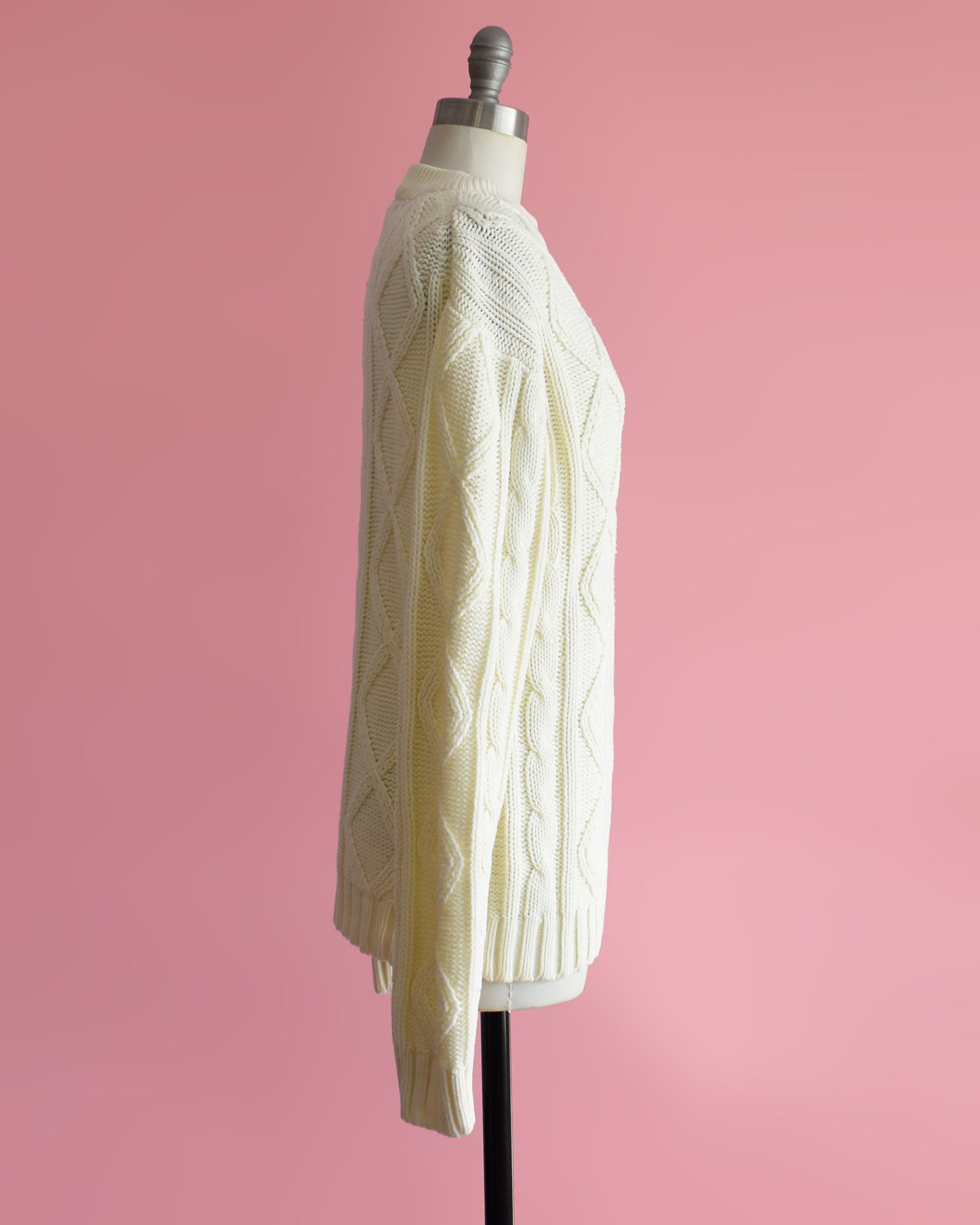 side view of a vintage 1970s cream cable knit sweater on a dress form against a pink background