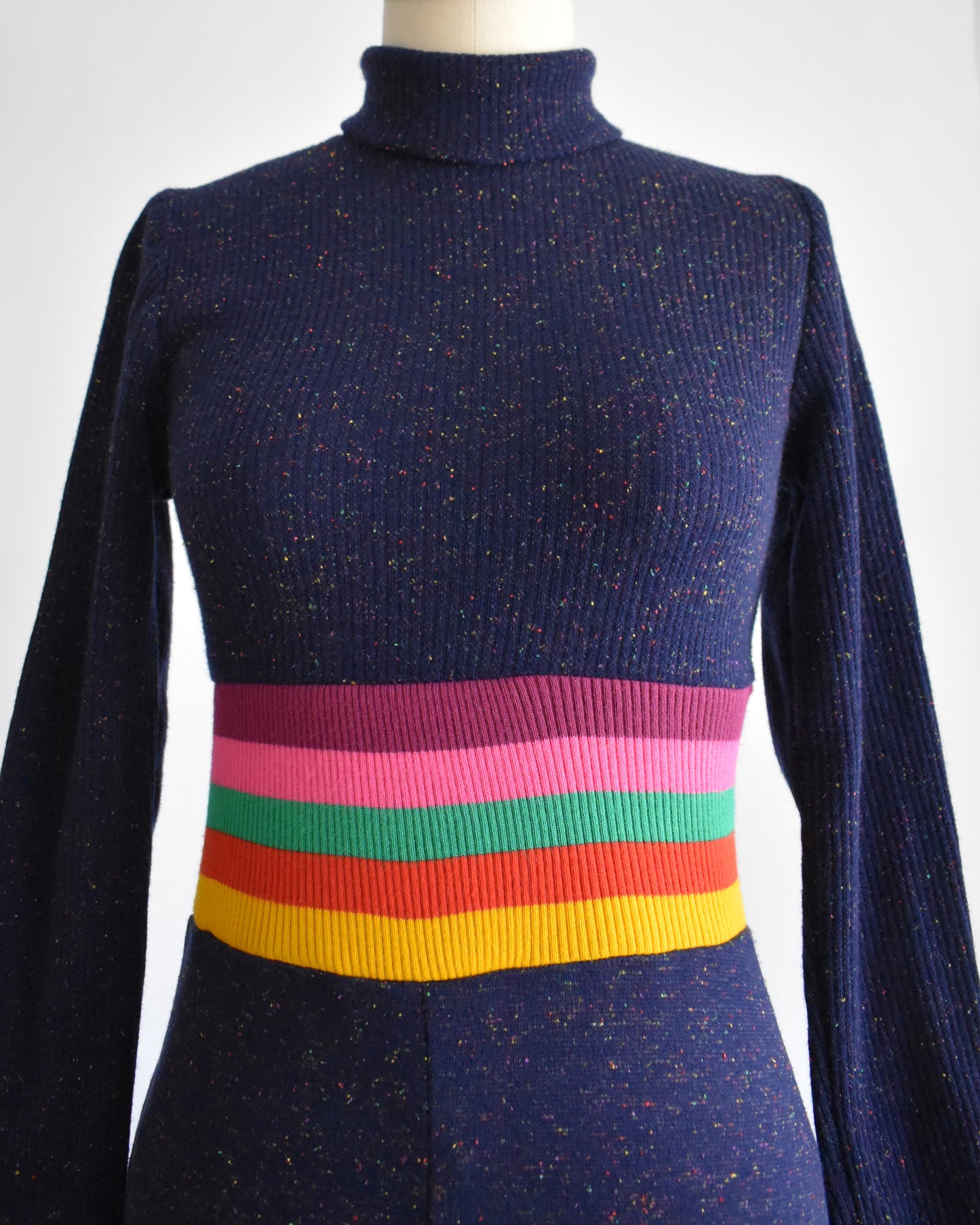 the front bodice of a vintage 1970s purple blue dress with a colorful striped waistband on a mannequin against a white background