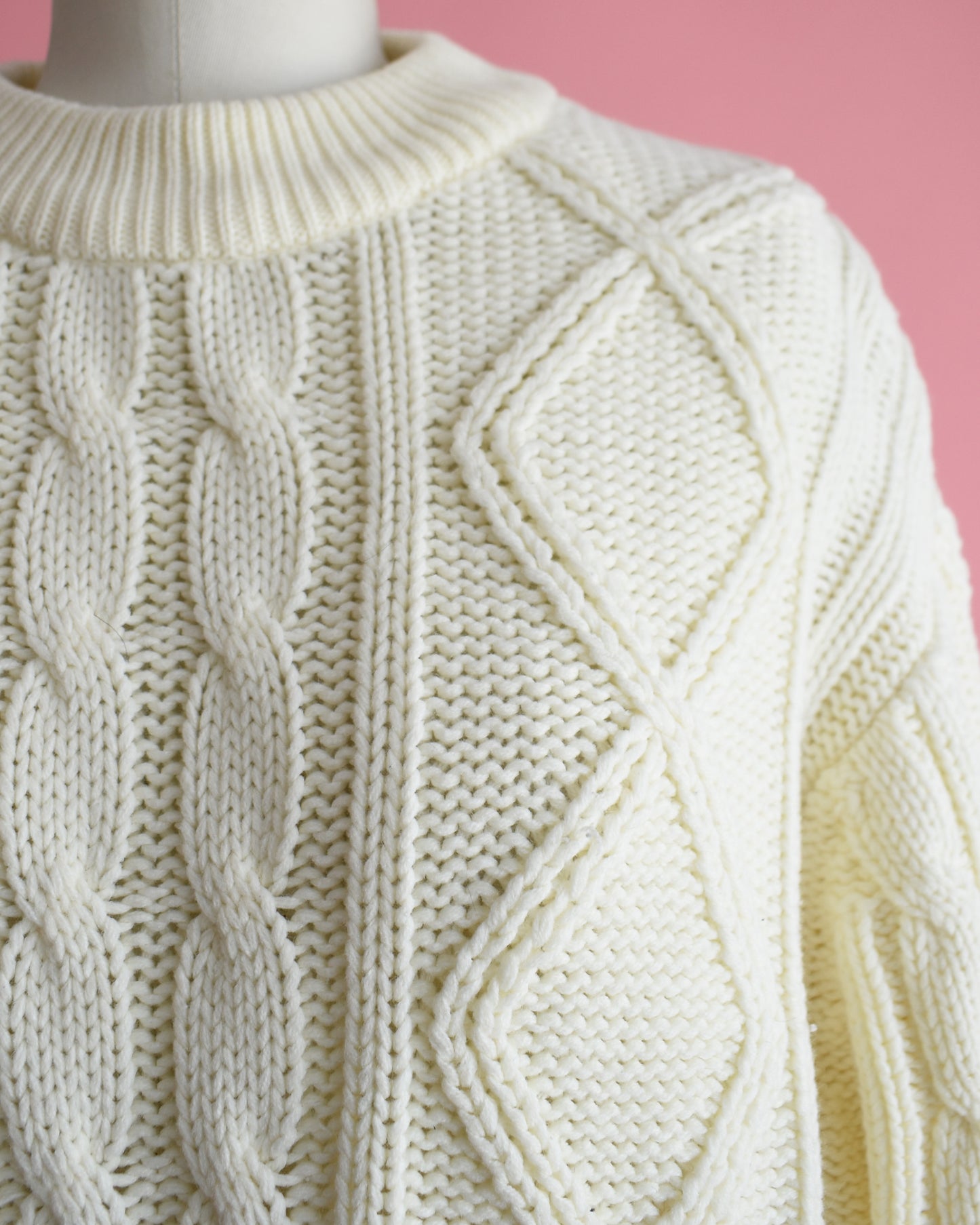 close up of the shoulder of a vintage 1970s cream cable knit sweater on a dress form against a pink background