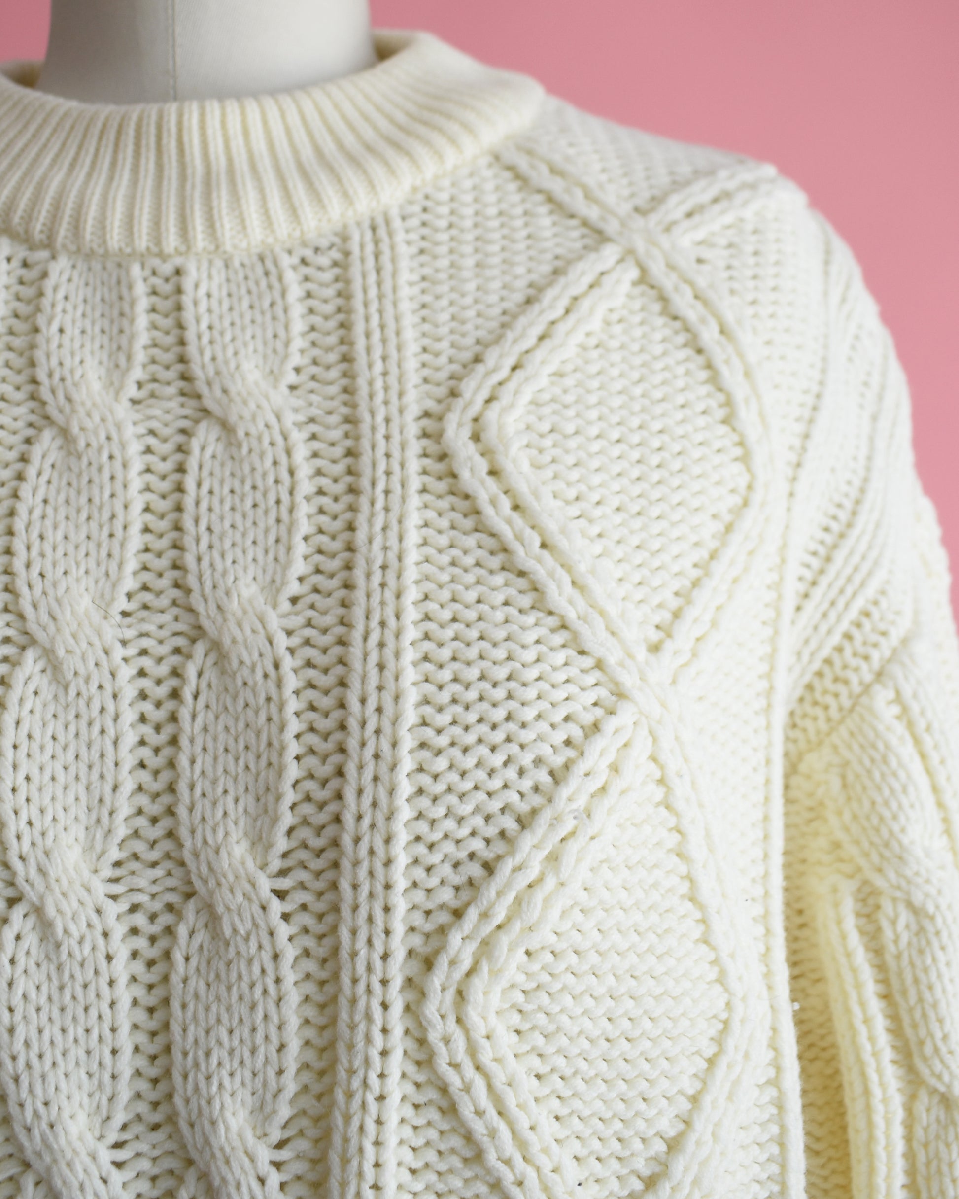 close up of the shoulder of a vintage 1970s cream cable knit sweater on a dress form against a pink background