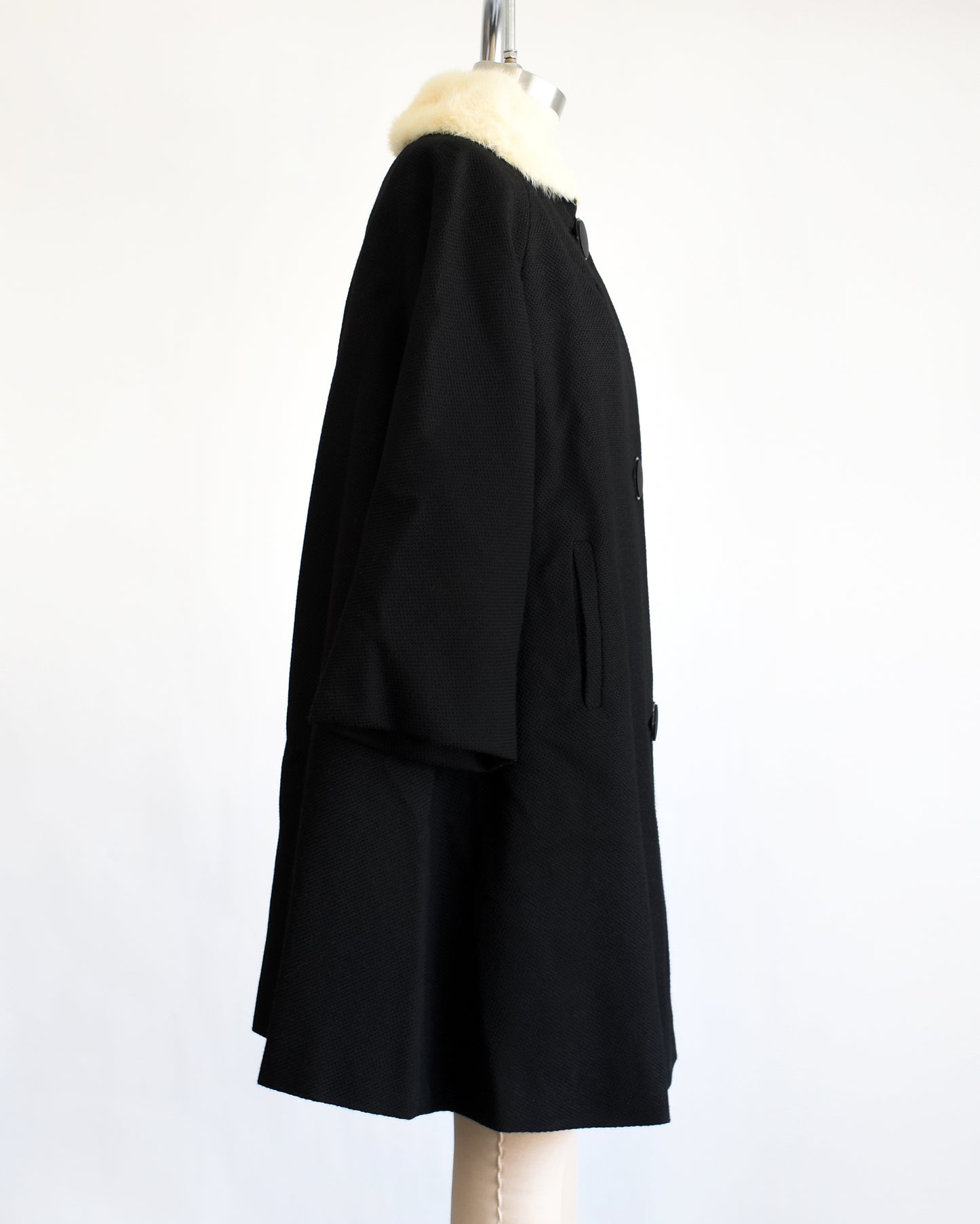 side view of a vintage 1950s black wool coat with fur collar on a mannequin against a white background