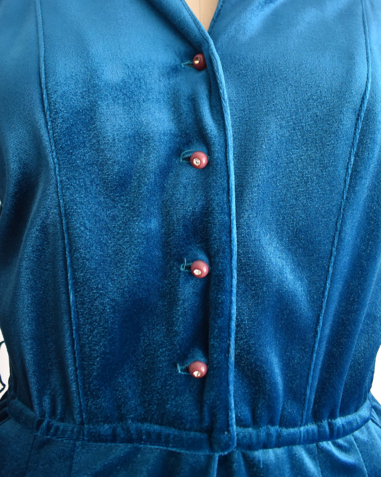 Close-up of a blue velvet jumpsuit with purple rhinestone buttons on a white background