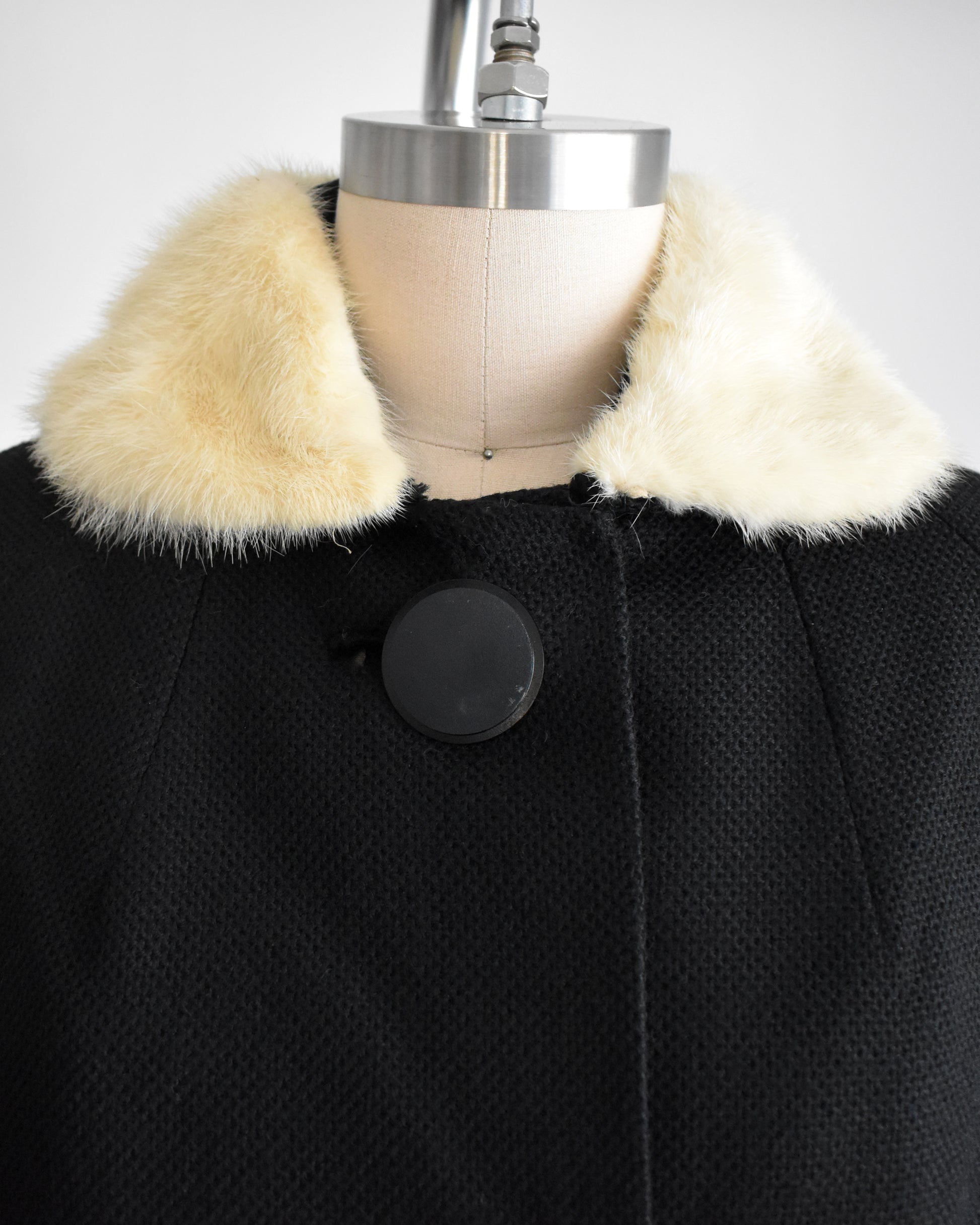 close up of the collar on a vintage 1950s black wool coat with fur collar on a mannequin against a white background