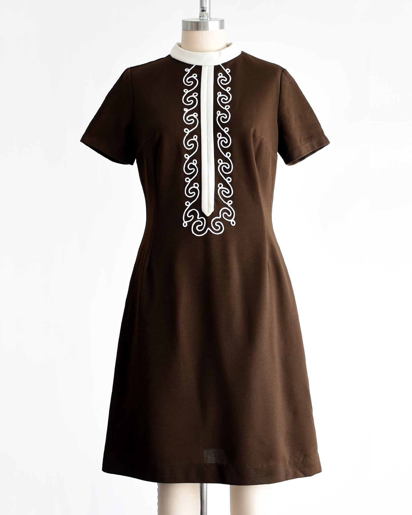 a vintage 1960s brown dress with white embroidery on a mannequin against a white background