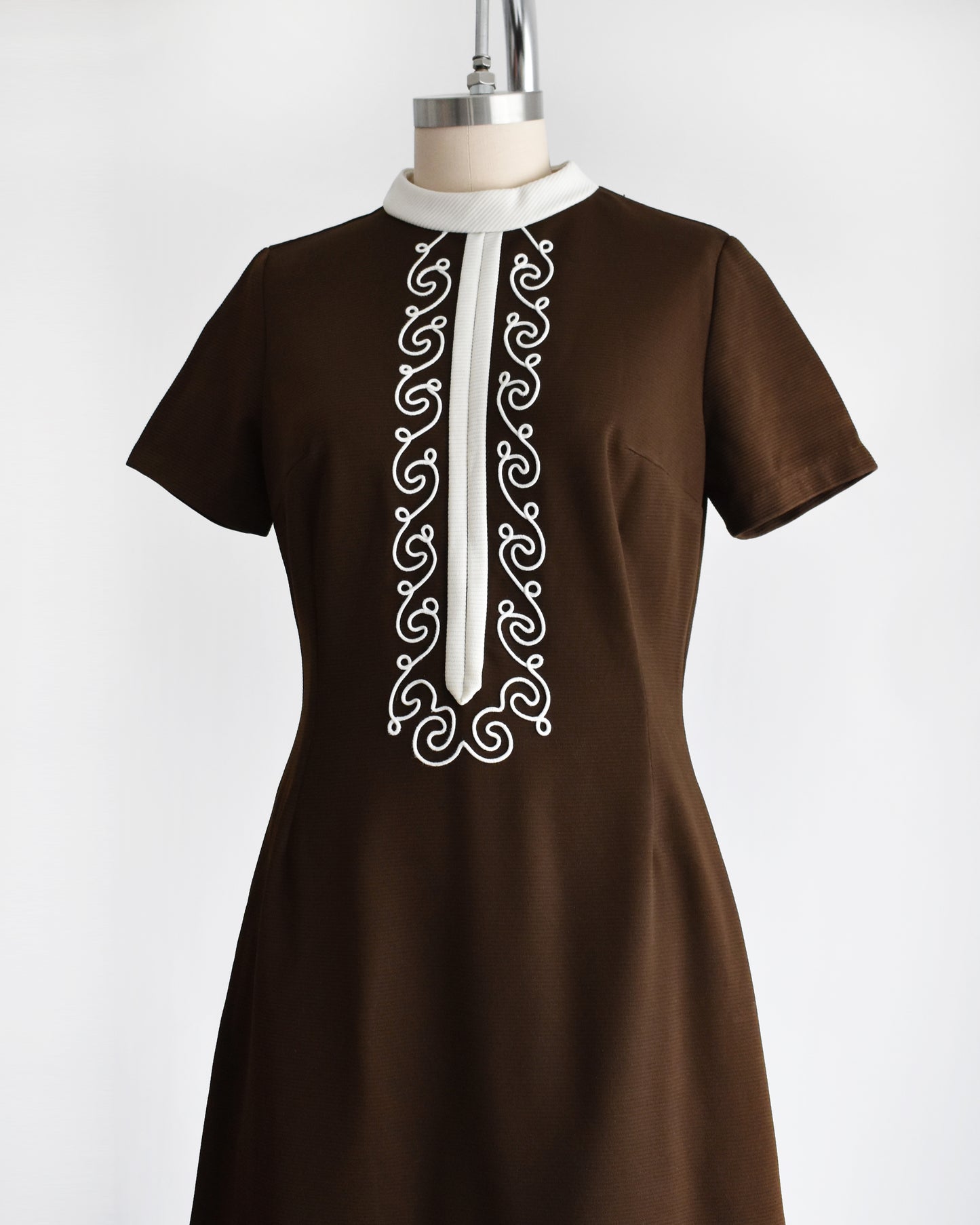 side front view of a vintage 1960s brown dress with white embroidery on a mannequin against a white background