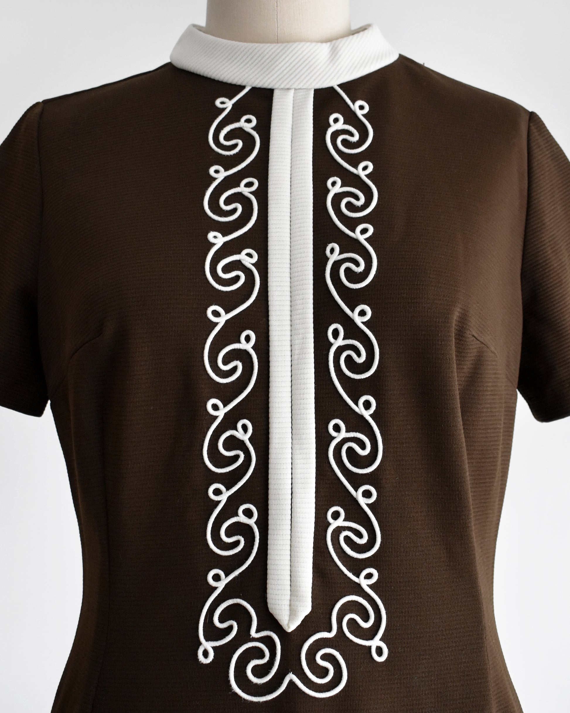 close up of the bodice of a vintage 1960s brown dress with white embroidery on a mannequin against a white background