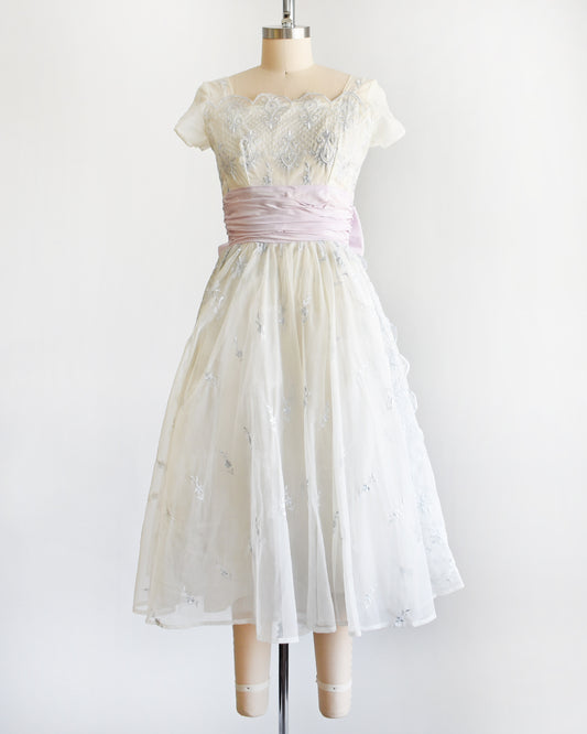 a vintage 1950s party dress that features icy blue floral embroidery on white chiffon fabric and a wide light purple waistband with a bow on the back