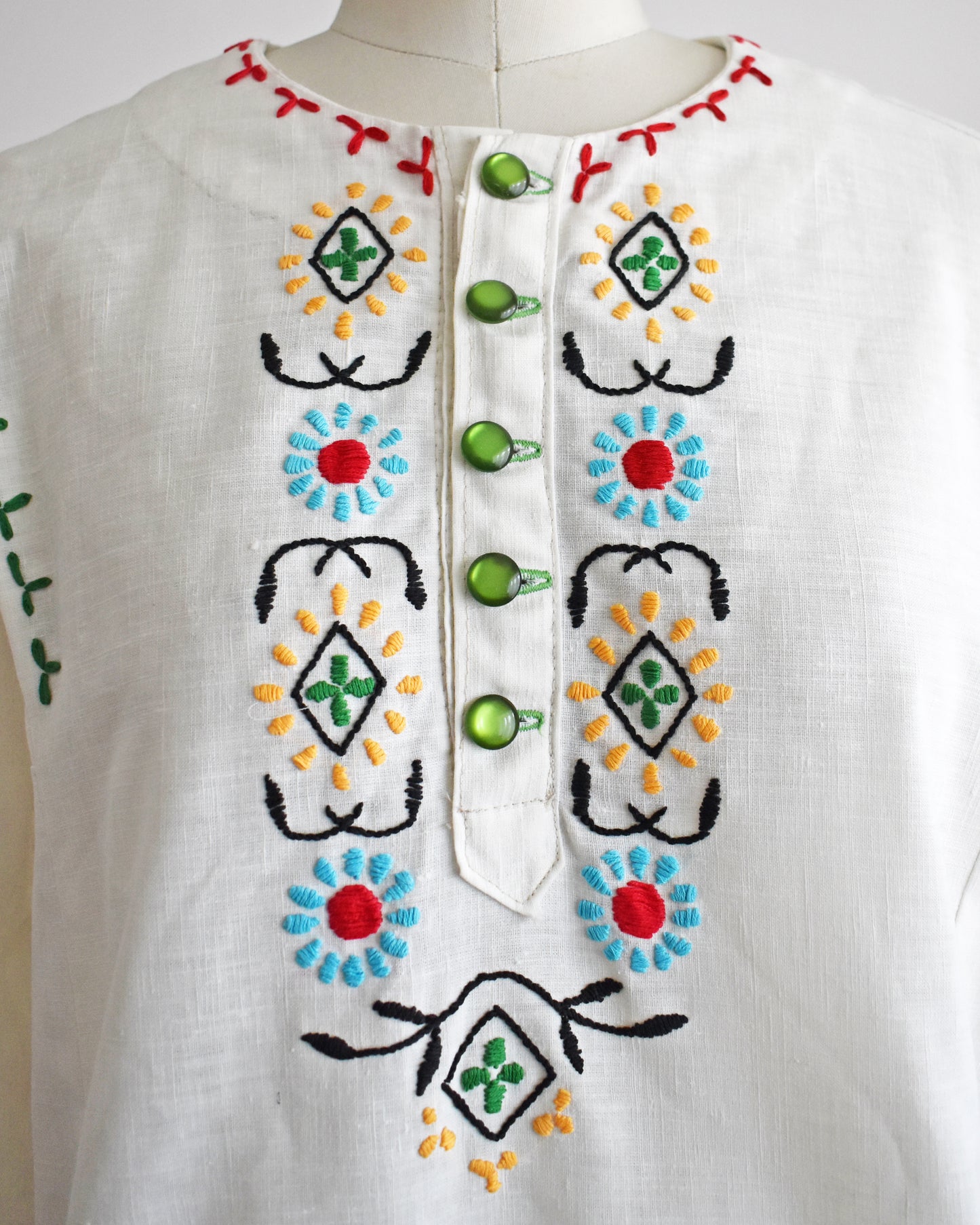 close up of the embroidery on a vintage 1970s white blouse with colorful embroidery on a mannequin against a white background
