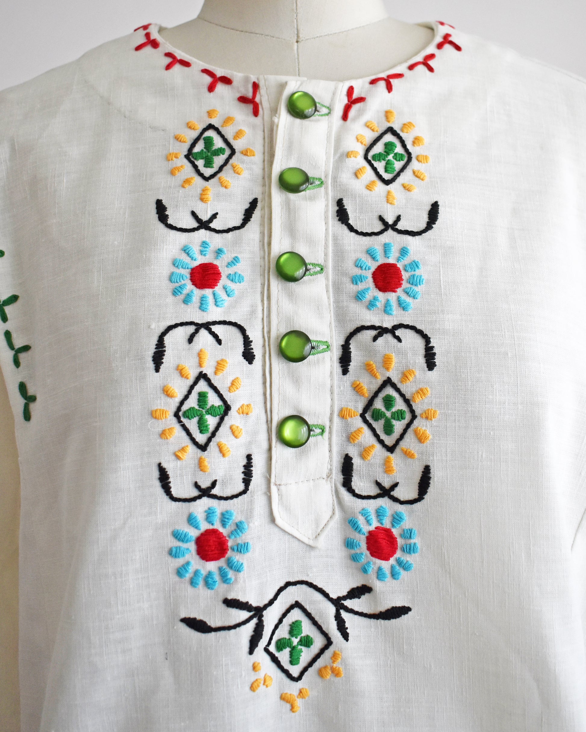 close up of the embroidery on a vintage 1970s white blouse with colorful embroidery on a mannequin against a white background