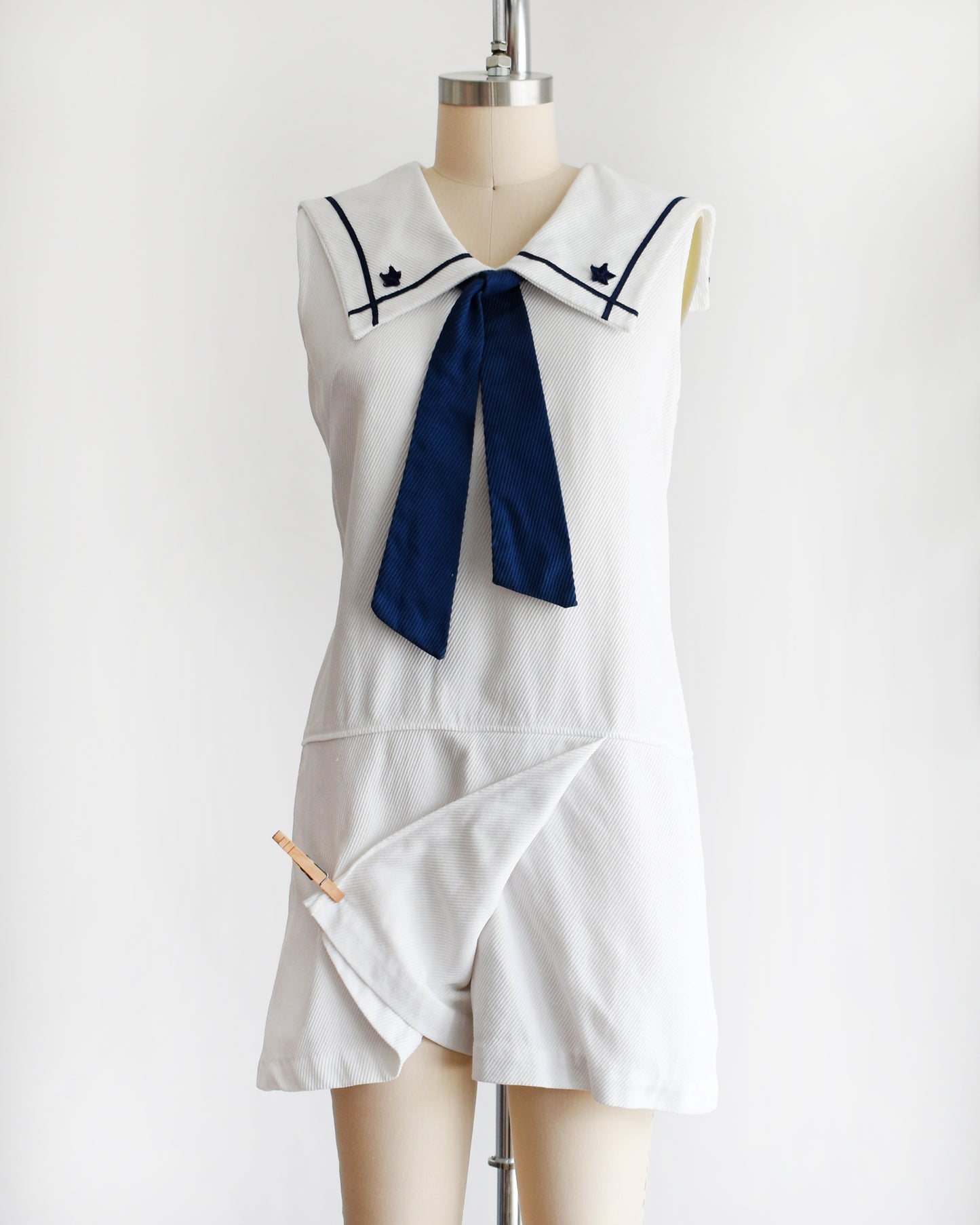 a vintage 1960s white romper dress with a navy blue bow on a mannequin against a white background. a clothespin has pulled back the skirt panel showing the shorts underneath