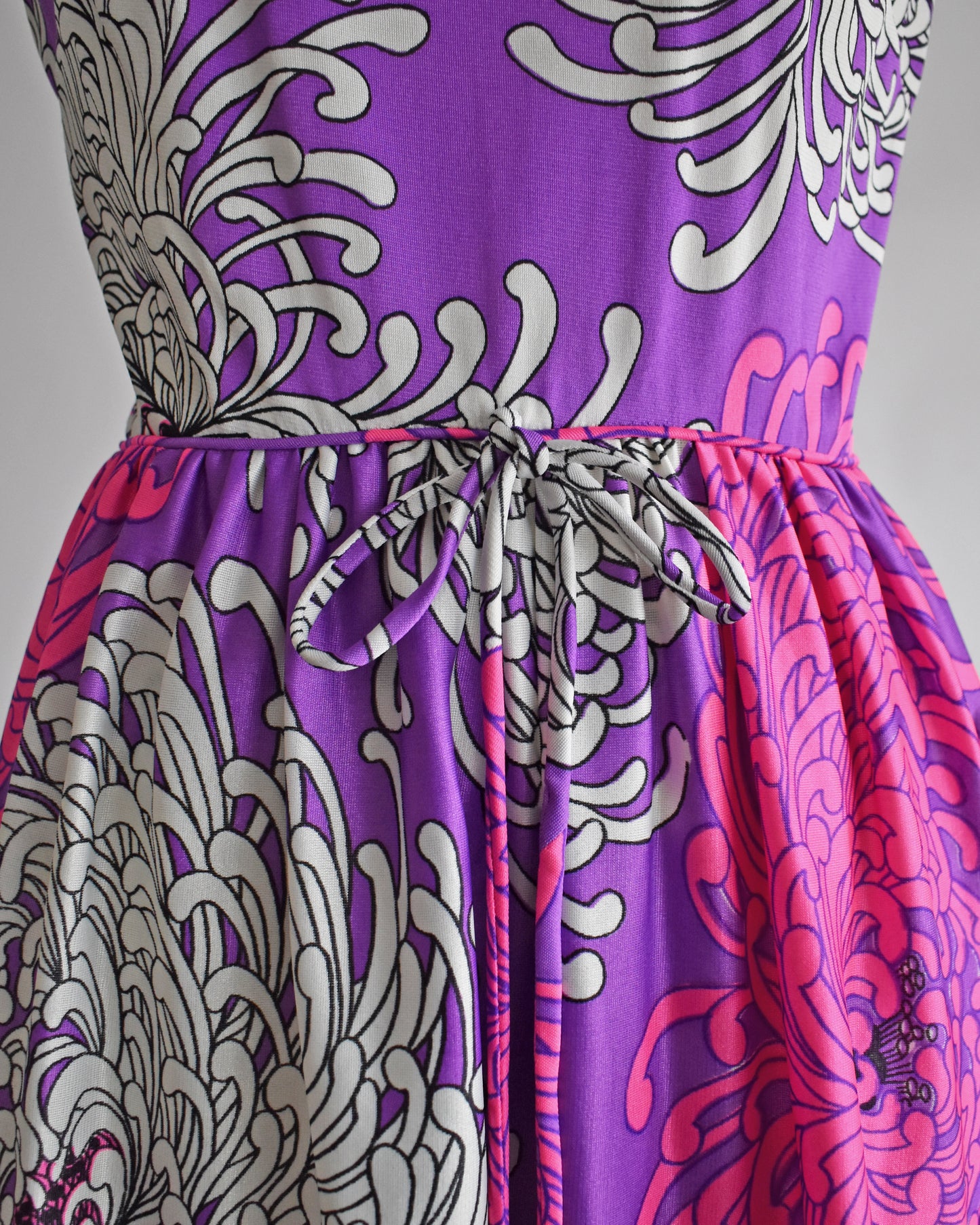 close up of the waist and bow on a vintage 1970s purple dress with floral patterns on a mannequin against a white background