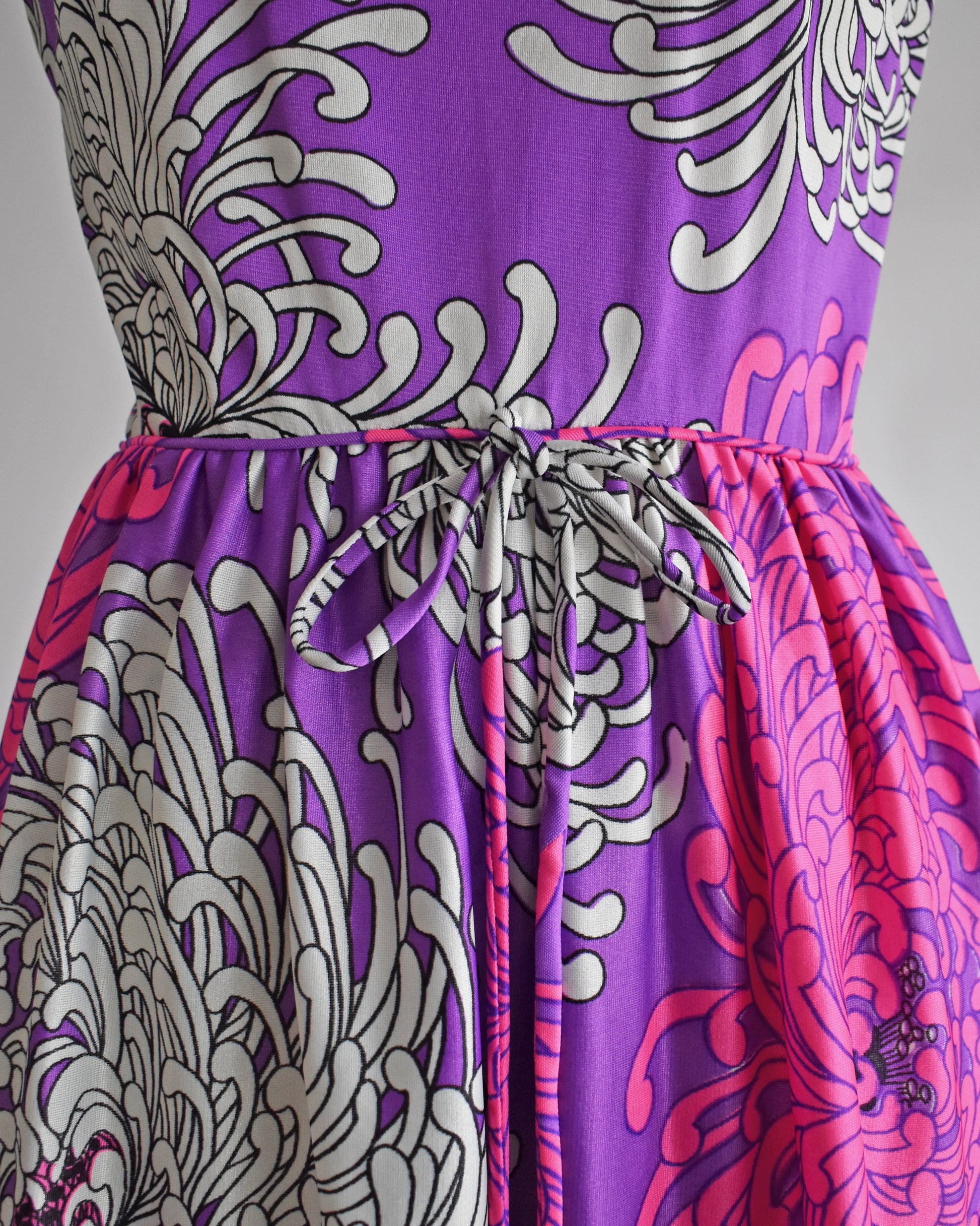 close up of the waist and bow on a vintage 1970s purple dress with floral patterns on a mannequin against a white background