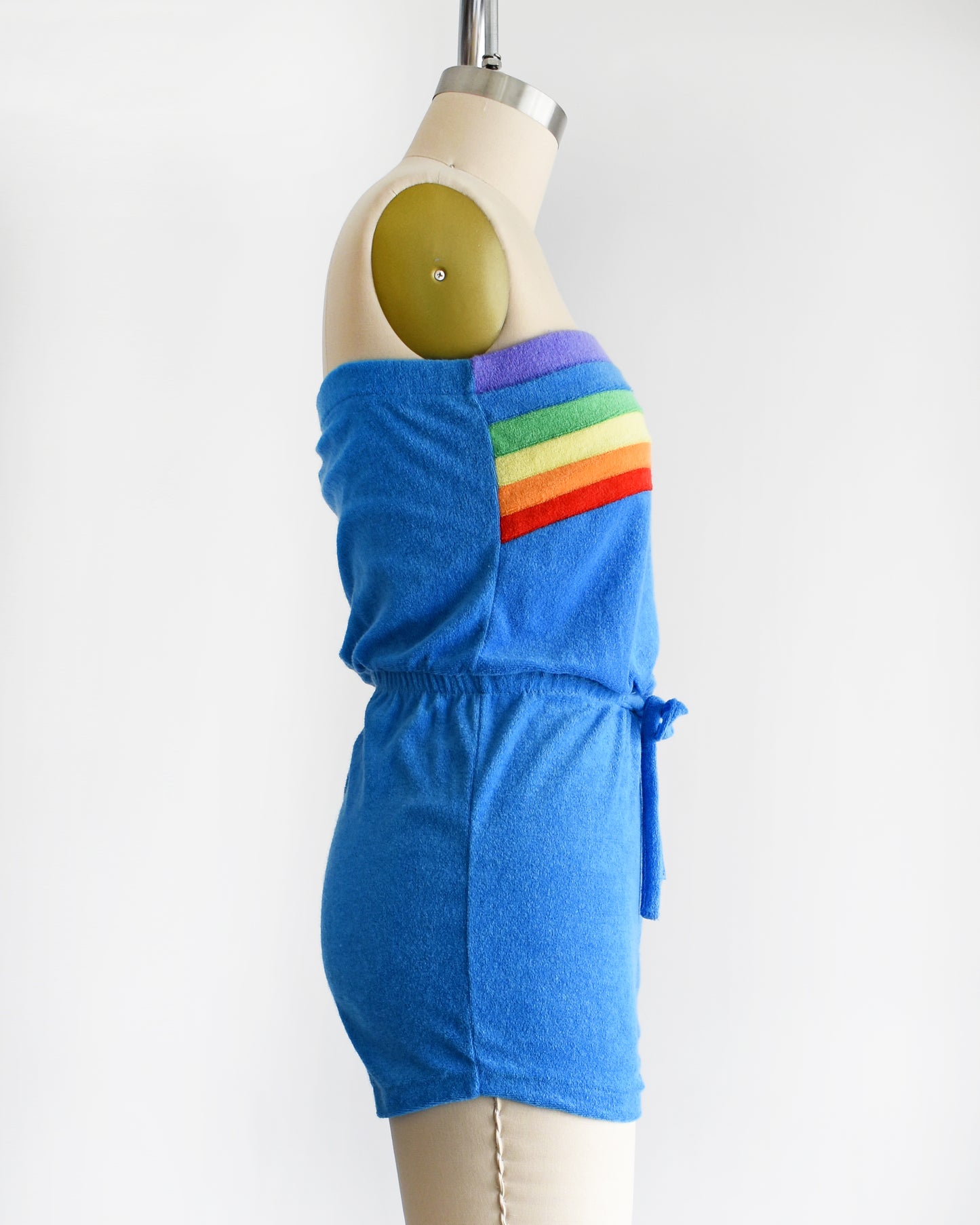 side view of a vintage late 1970s early 1980s blue terry cloth romper that has a rainbow stripe on the top 