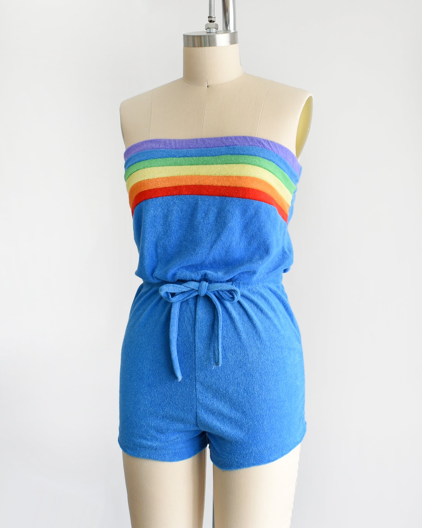 side front view of a vintage late 1970s early 1980s blue terry cloth romper that has a rainbow stripe on the top 