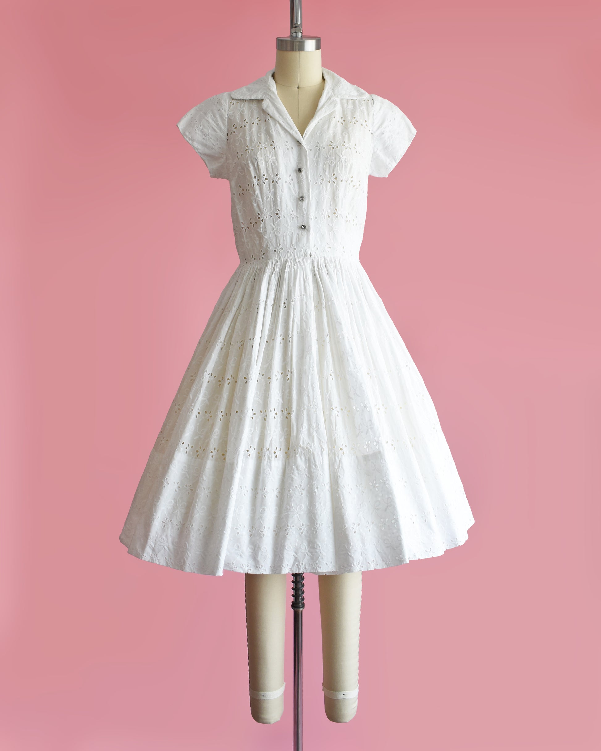a vintage 1950s white eyelet dress with rhinestone buttons on a mannequin against a pink background