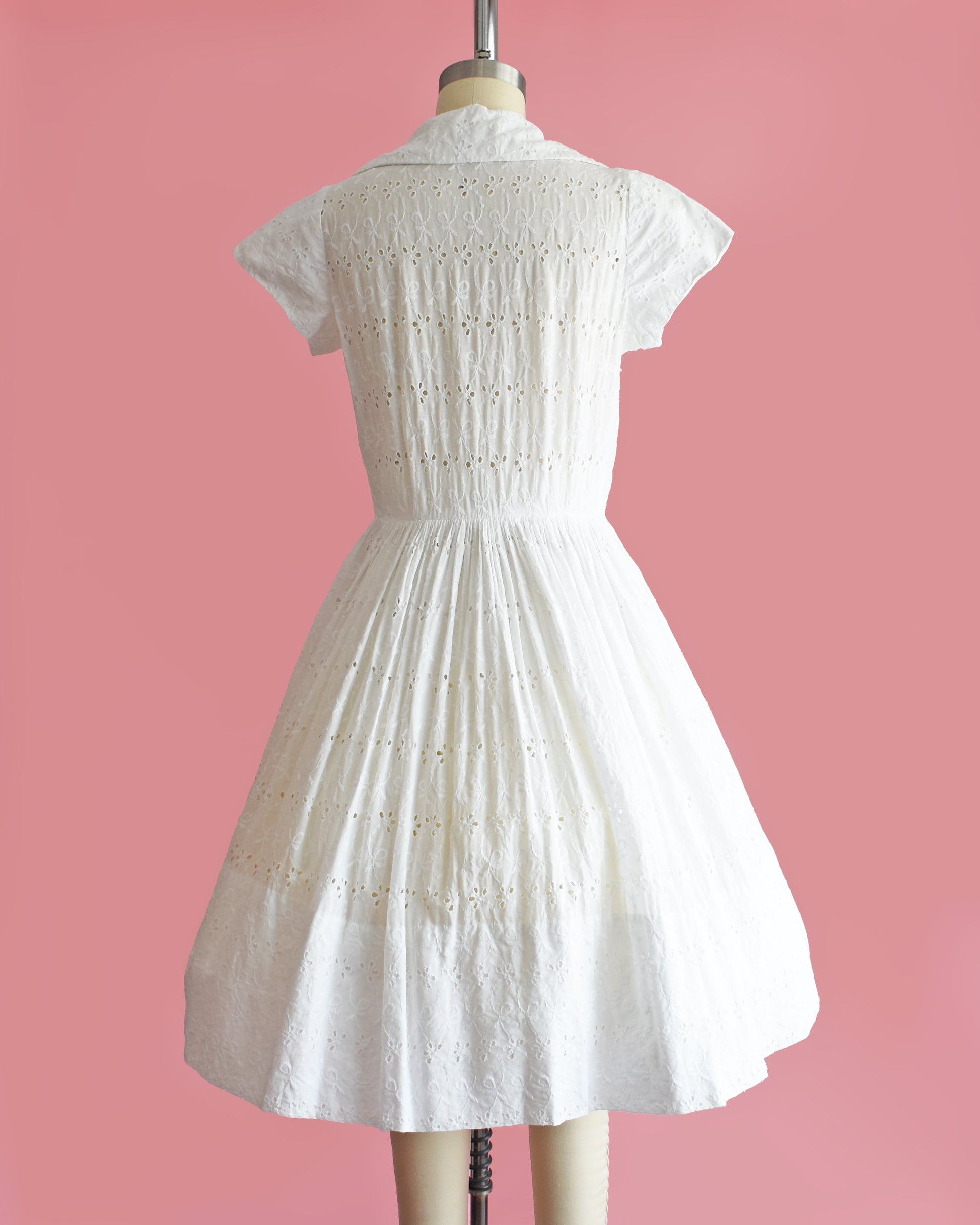 back view of a vintage 1950s white eyelet dress with rhinestone buttons on a mannequin against a pink background