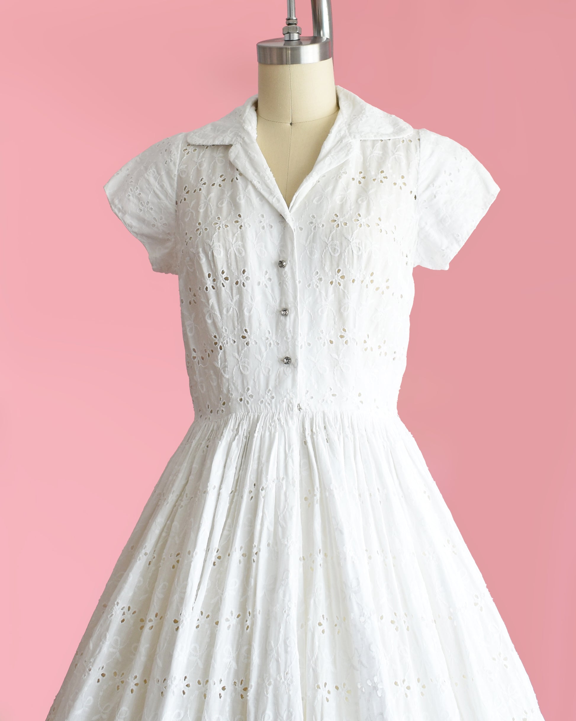 side front view of a vintage 1950s white eyelet dress with rhinestone buttons on a mannequin against a pink background