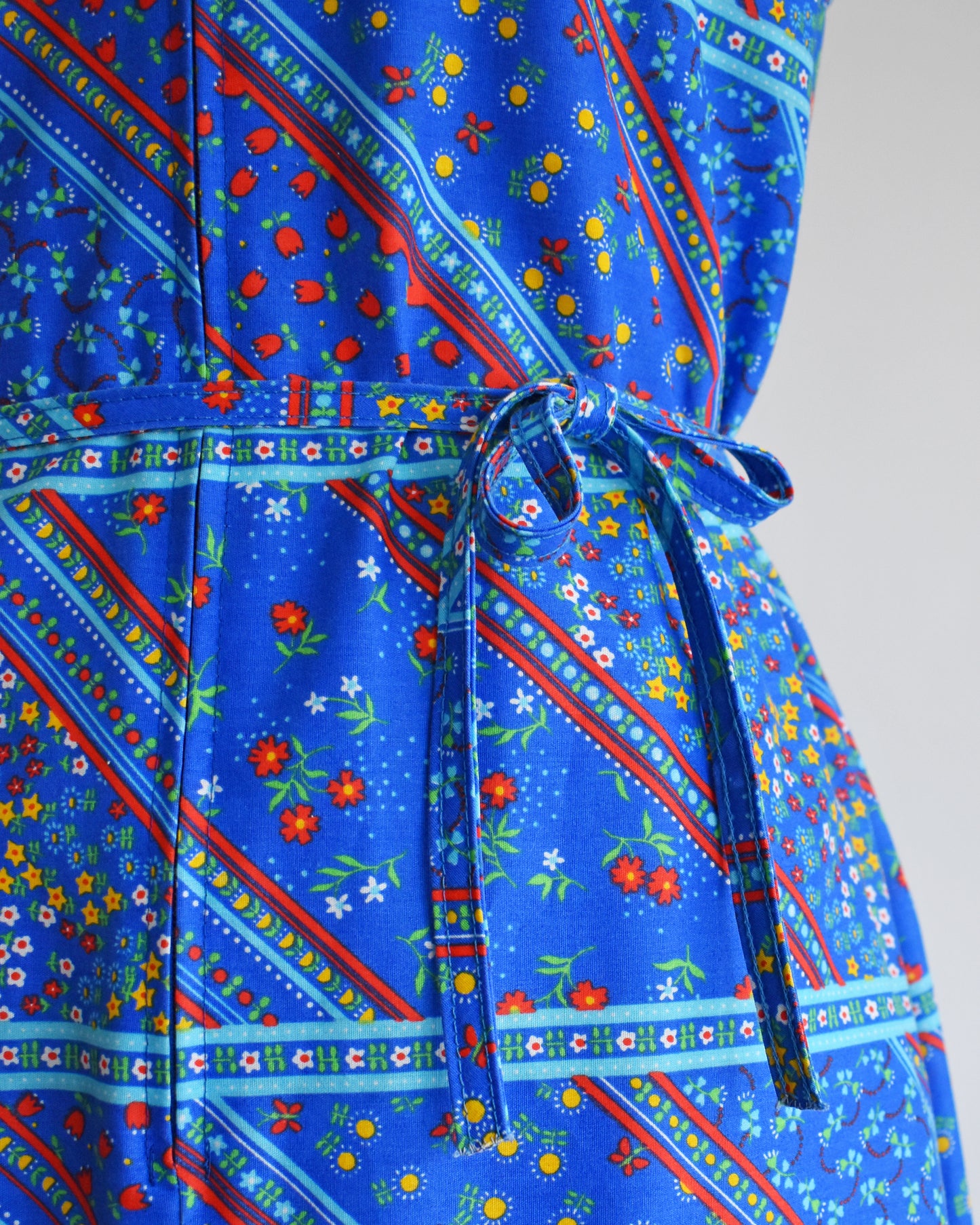 close up of the tie belt on the waist of a vintage 1970s blue floral dress on a mannequin against a white background
