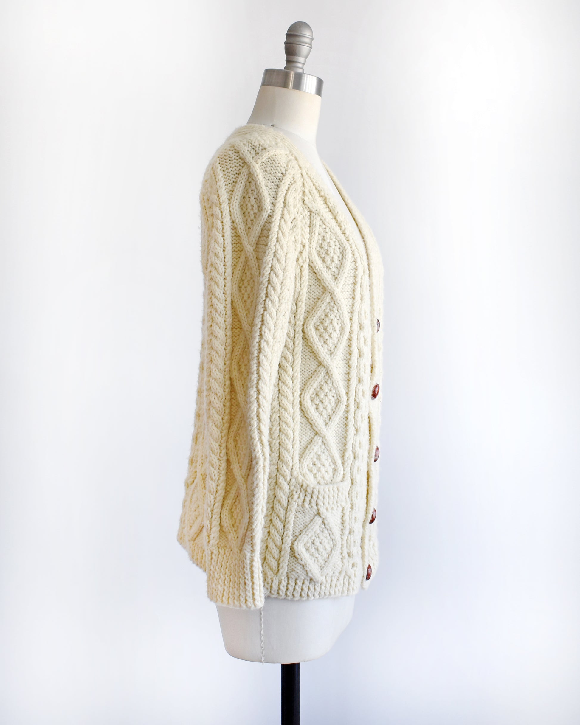 side view of a vintage 1970s/1980s cream-colored cable knit cardigan on a mannequin against a white background