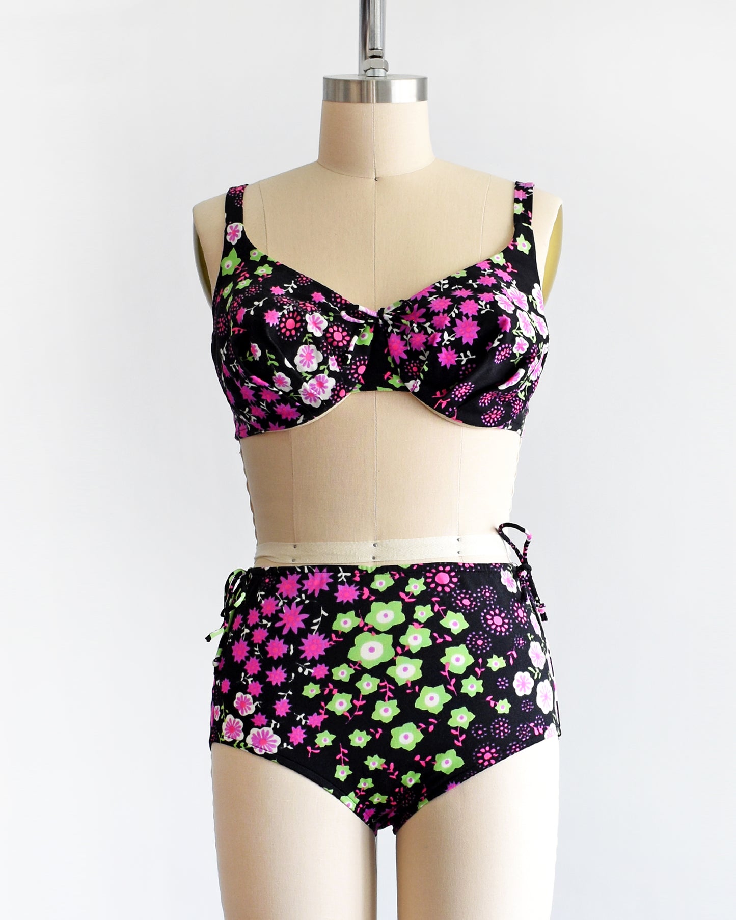 a vintage 1960s two piece bikini set that is black and has a bright pink, purple, green, and white flower power print on a dress form.