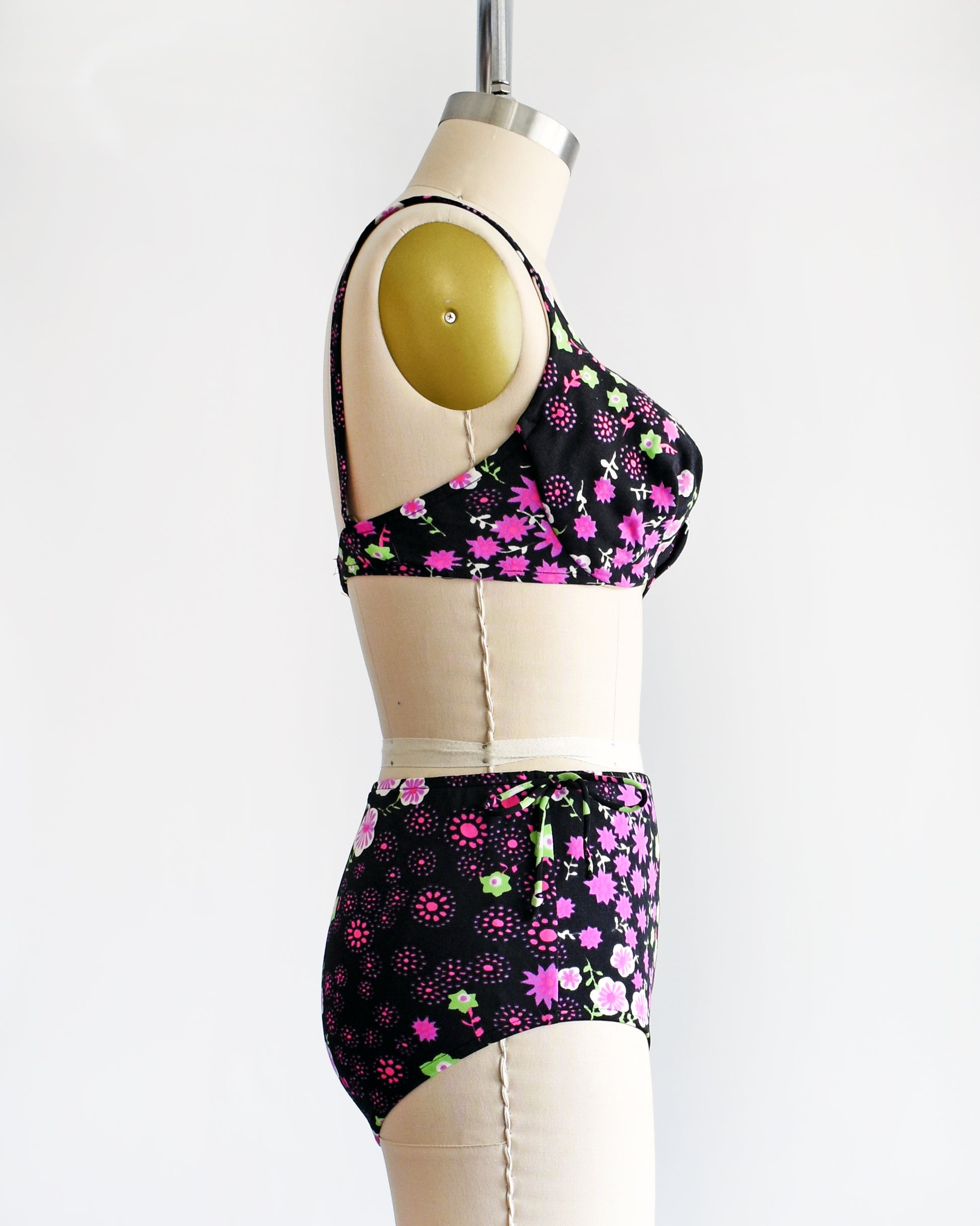 side view of a vintage 1960s two piece bikini set that is black and has a bright pink, purple, green, and white flower power print on a dress form.