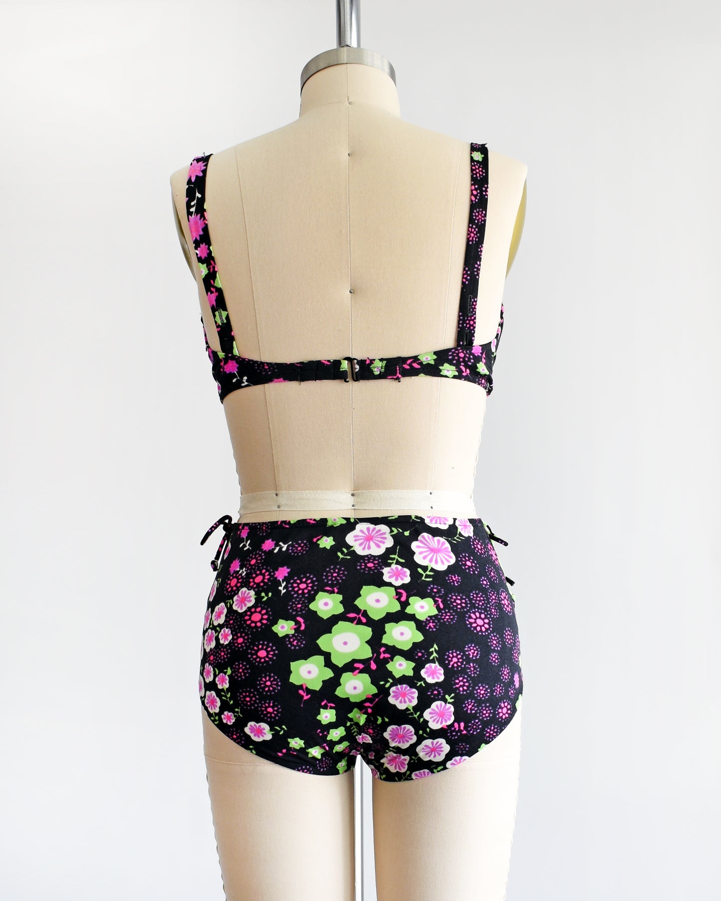 back view of a vintage 1960s two piece bikini set that is black and has a bright pink, purple, green, and white flower power print on a dress form.