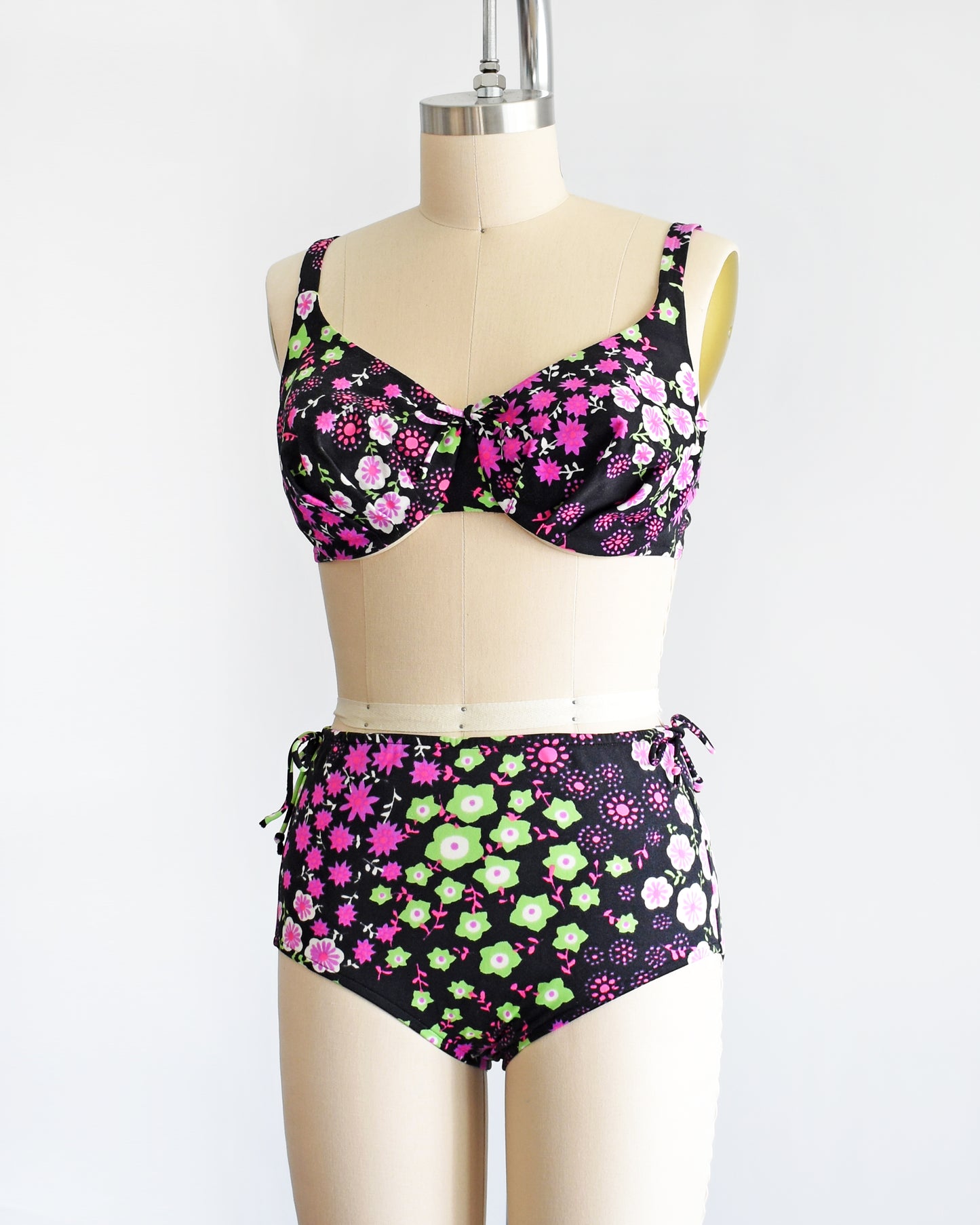 side front view of a vintage 1960s two piece bikini set that is black and has a bright pink, purple, green, and white flower power print on a dress form.