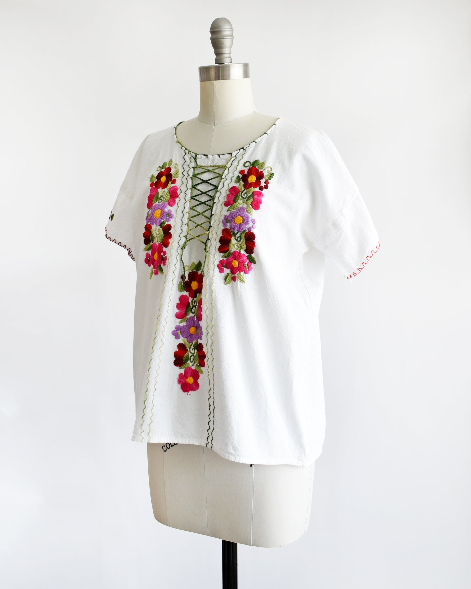 side front view of a vintage 1970s white blouse with colorful floral embroidery on a mannequin against a white background