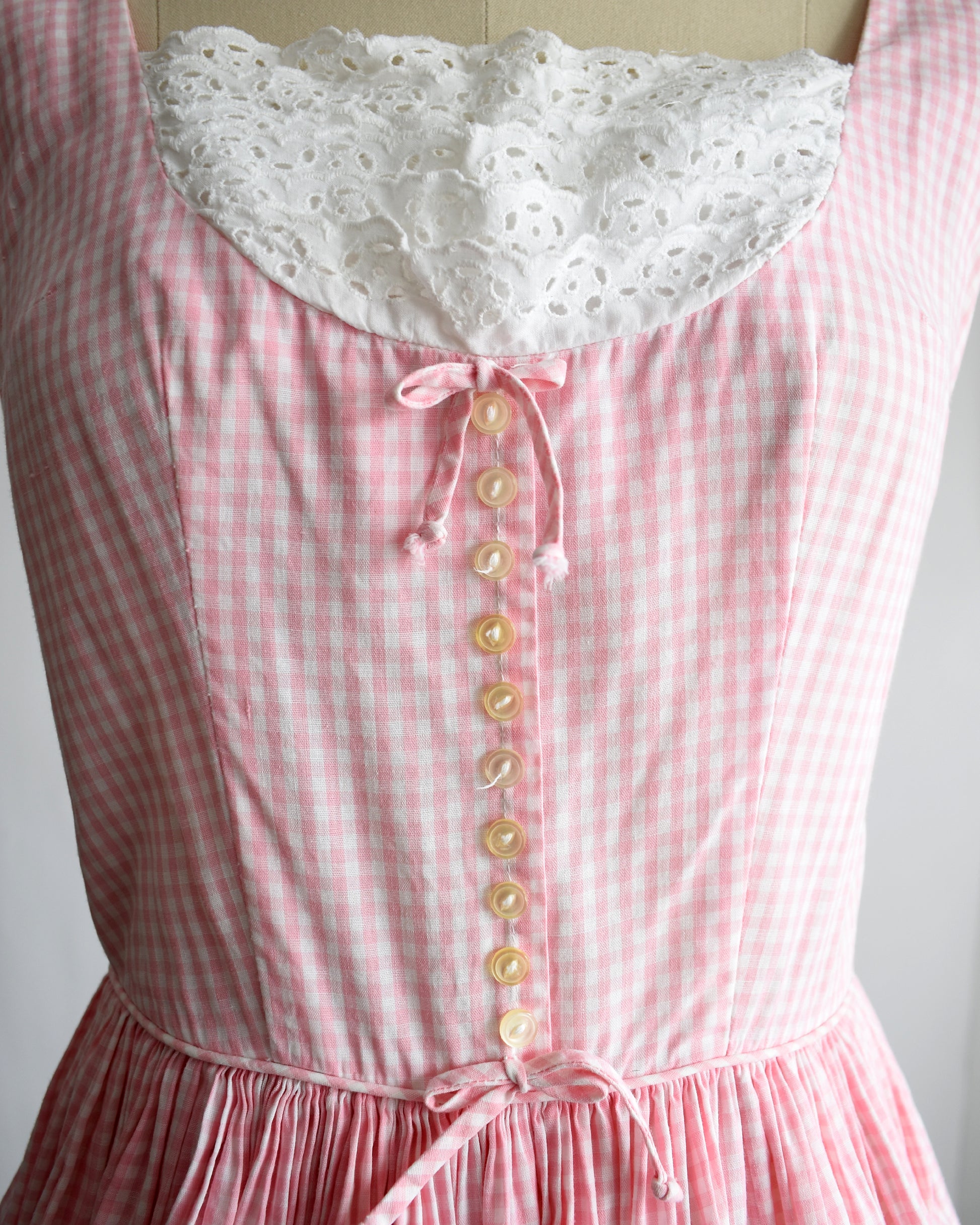 close up of a vintage 1950s pink and white gingham dress with a white lace bib on a mannequin against a white background