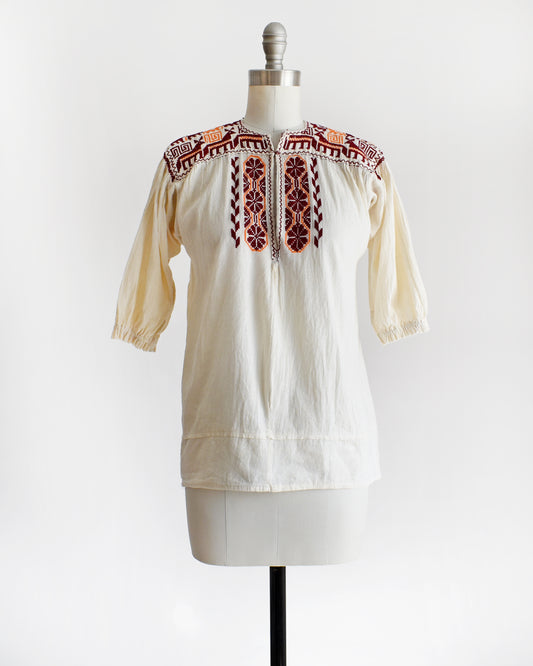 A vintage 60s cream crinkled cotton top with a burgundy and orange embroidered floral and what looks like llamas motif across the shoulders and down the front. The top is on a dress form.