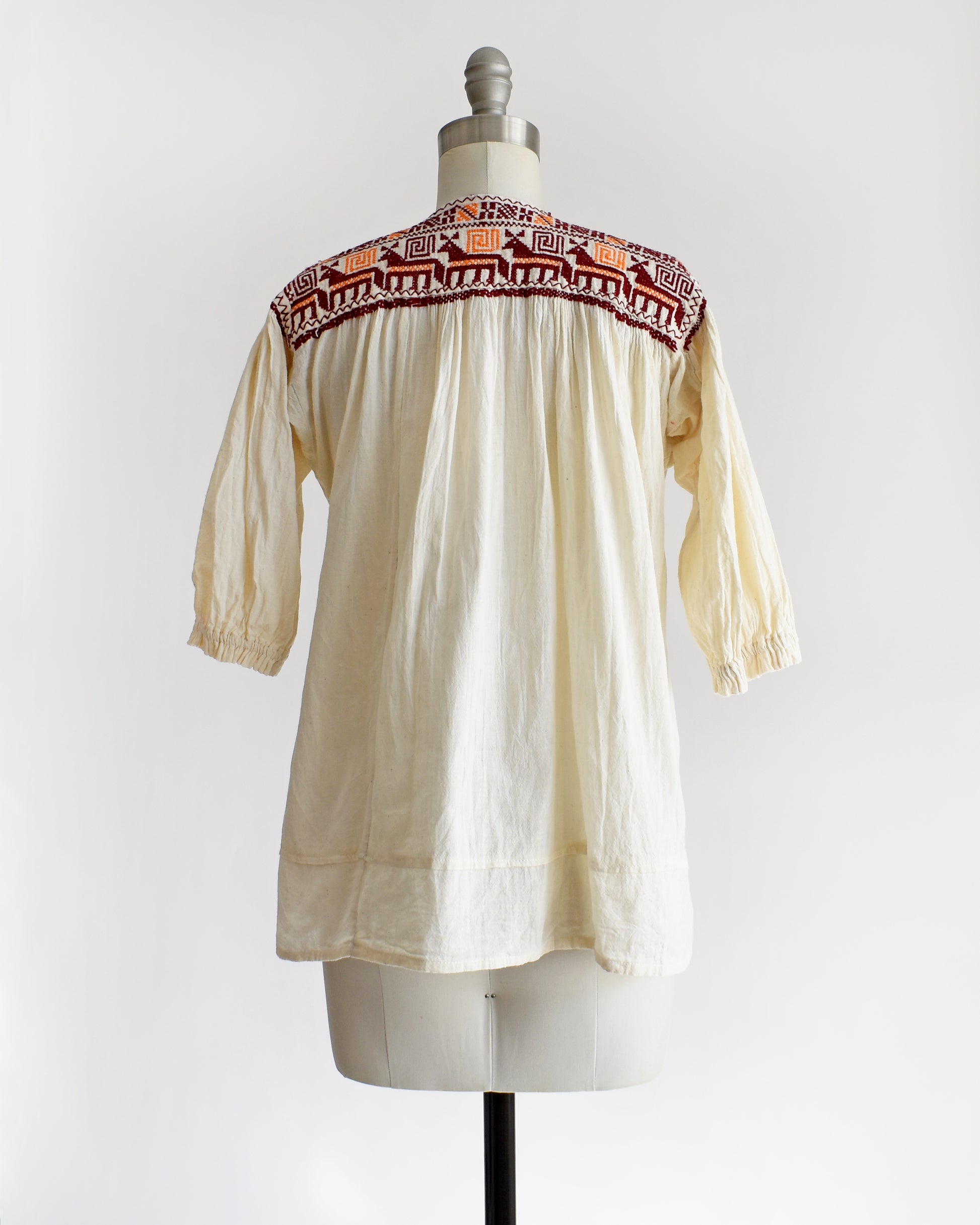 Back view of a vintage 60s cream crinkled cotton top with a burgundy and orange embroidered floral and what looks like llamas motif across the shoulders and down the front. The top is on a dress form.