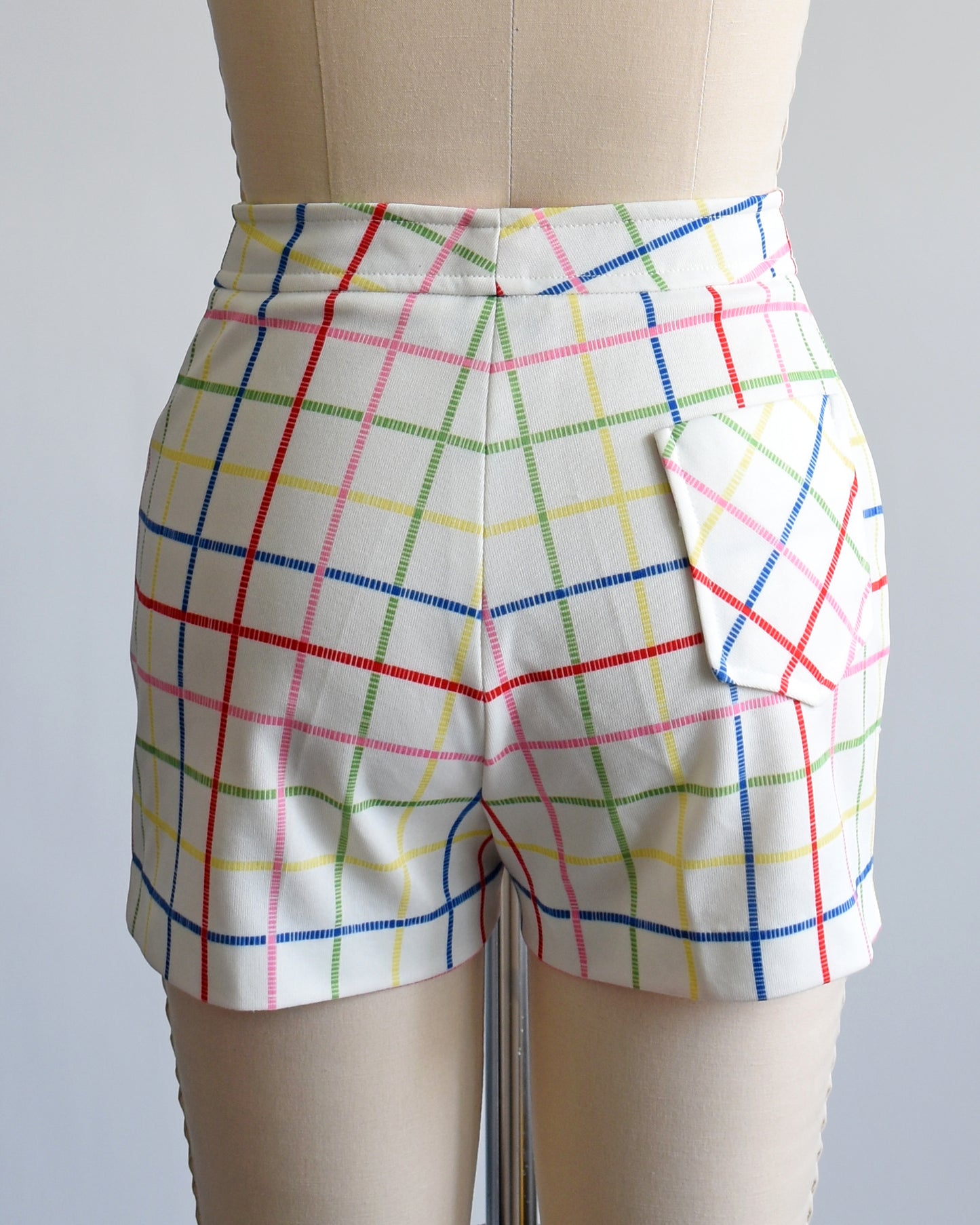 back view of a pair of vintage 1970s white shorts with rainbow plaid stripes on a dress form. there is a pocket on the back right of the shorts