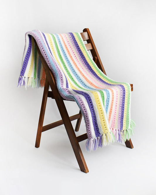 A white with dark and light purple, pink, blue, green, yellow, cream, and orange striped fringed blanket on a chair.