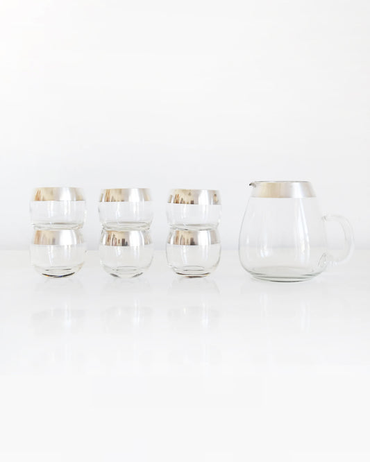 A vintage Dorothy Thorpe cocktail set that has a set of six silver rimmed rounded roly poly glasses with a matching silver rim pitcher with handle. The glasses are stacked creating a row of three