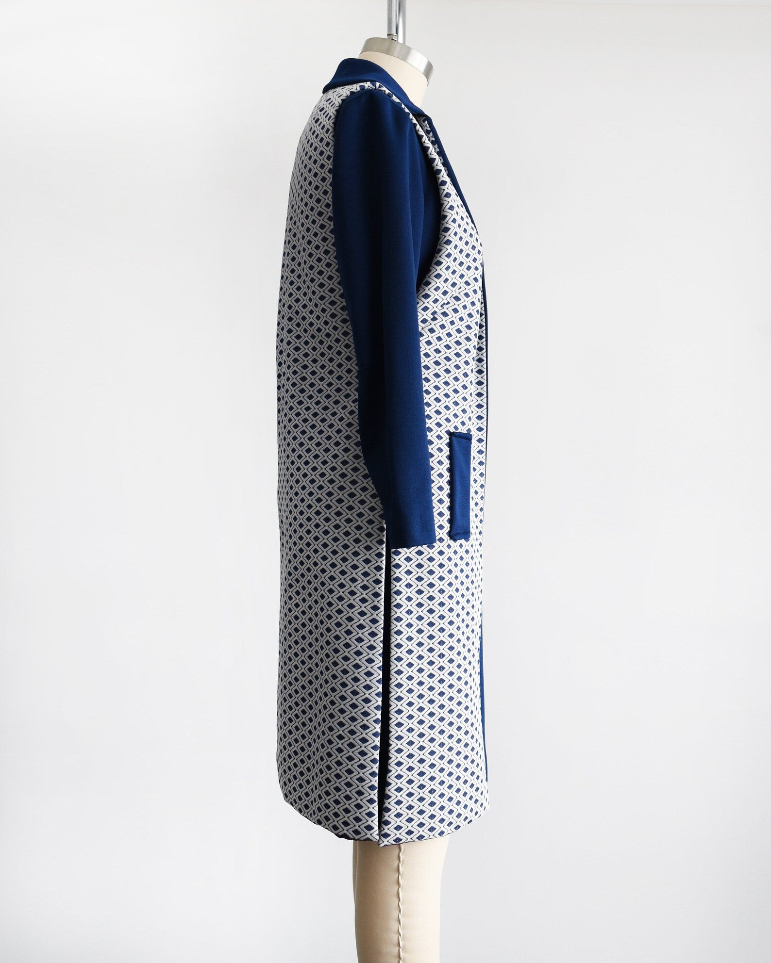 Side view of a vintage 60s/70s two piece dress set that features a long sleeve navy dress with a white and navy diamond print long vest with side slits. Dress is modeled on a dress form.