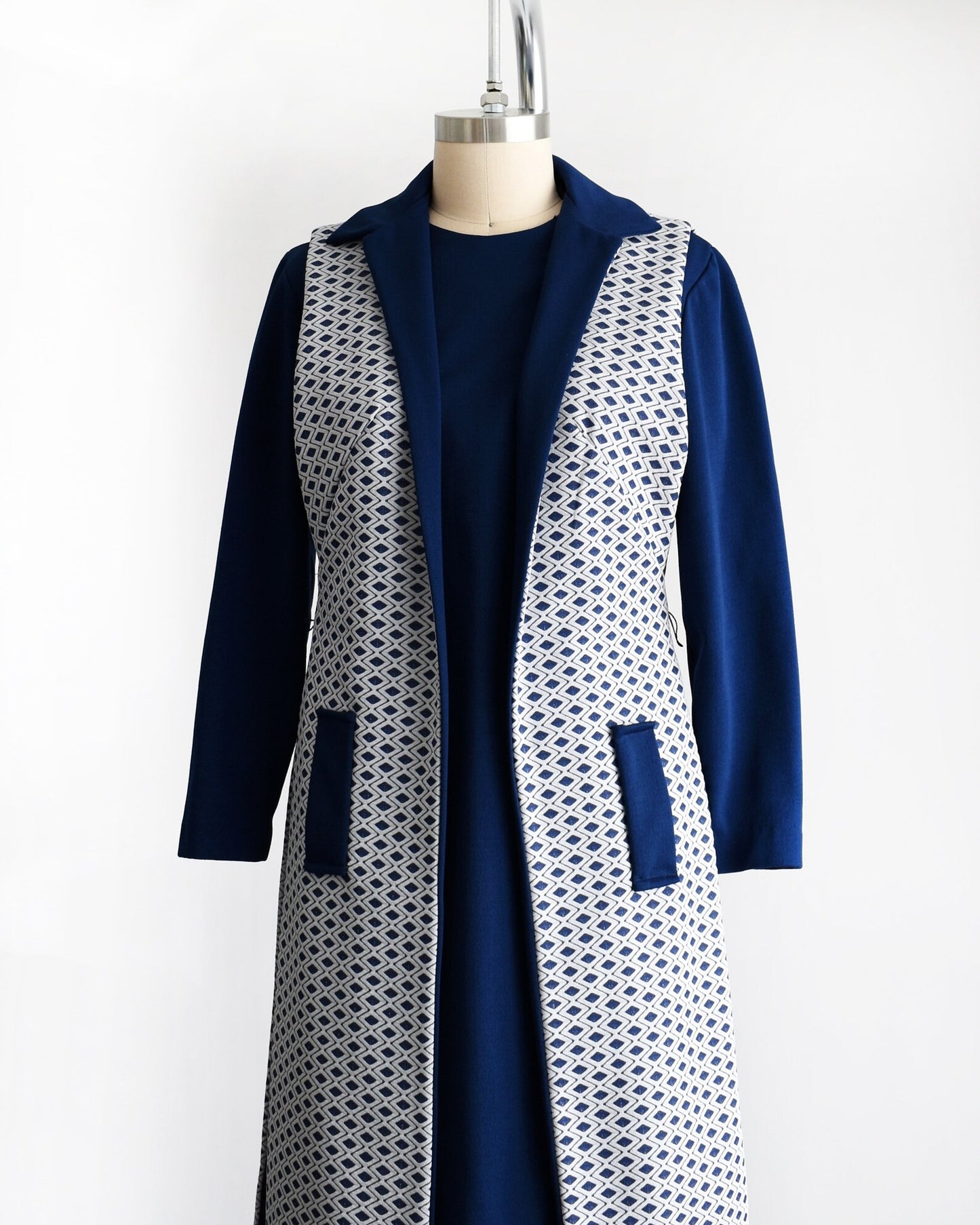 Side front view of a vintage 60s/70s two piece dress set that features a long sleeve navy dress with a white and navy diamond print long vest. Dress is modeled on a dress form.