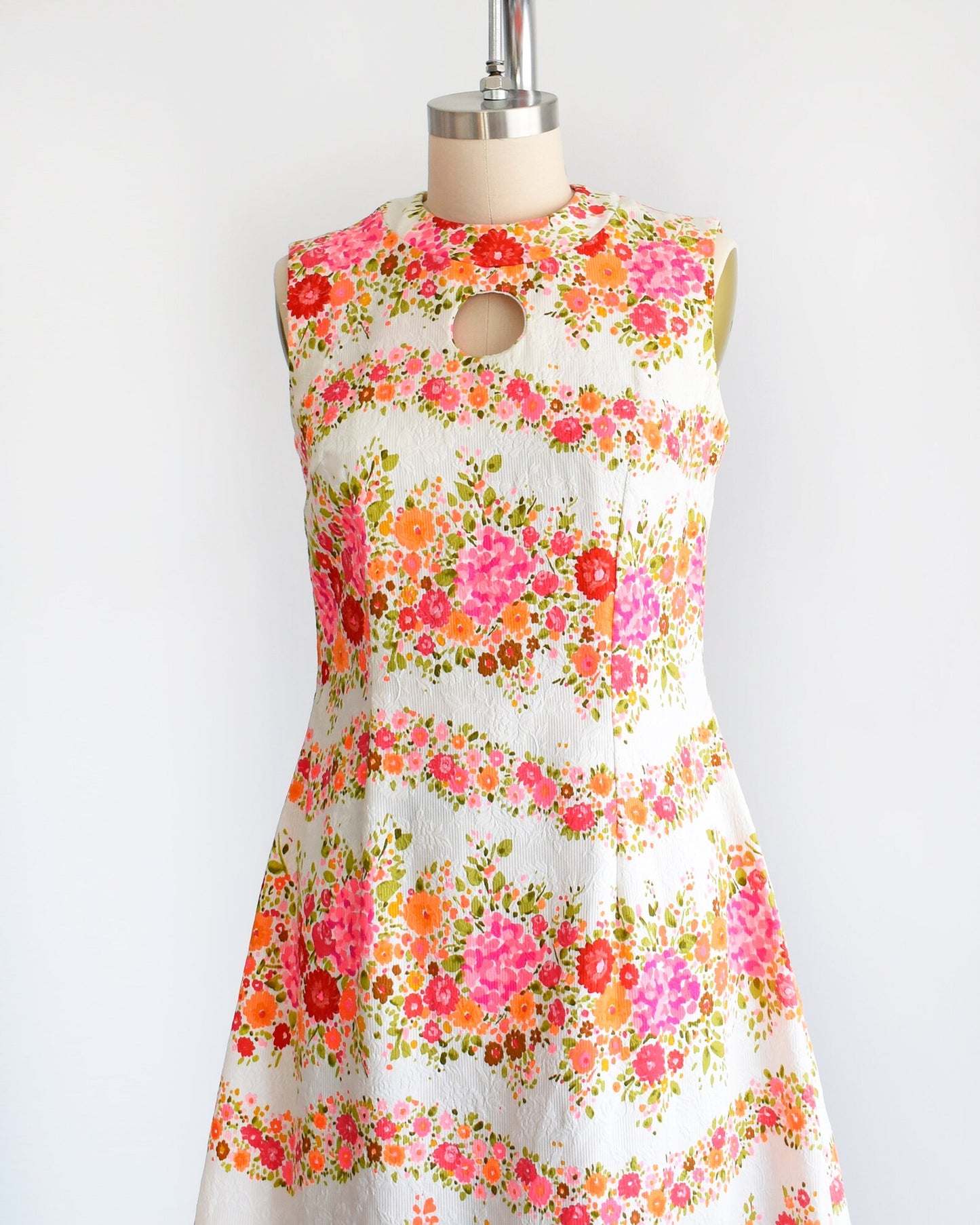 Side front view of a vintage 60s/70s white dress that has a vibrant pink, orange, and green floral print in wavy horizontal stripes going down the dress. There is a circle cut out under the collar. The dress is on a dress form.